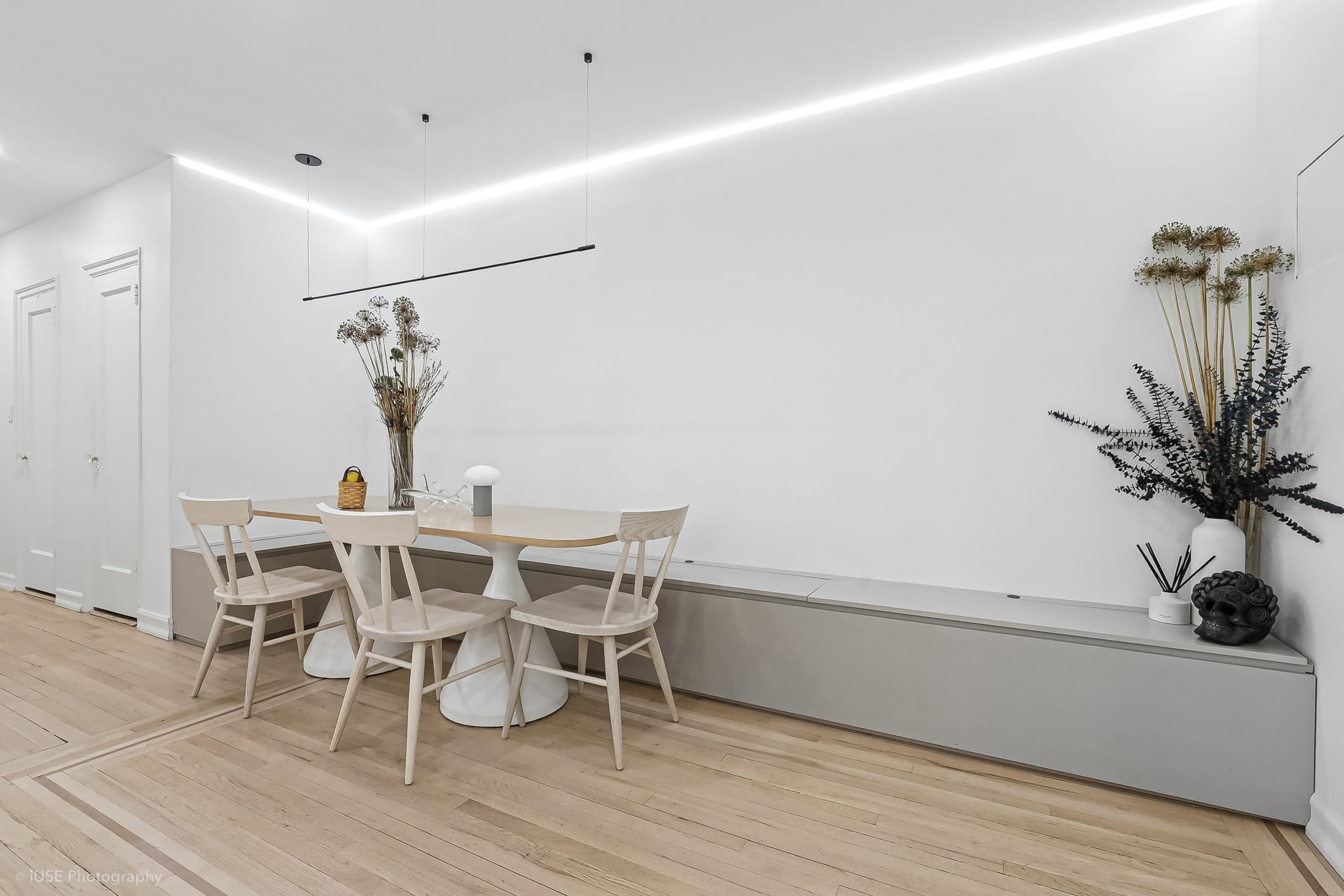 A clean, minimalist dining area features a wooden table with four chairs, a long storage unit against the wall, and decorative plants in vases.