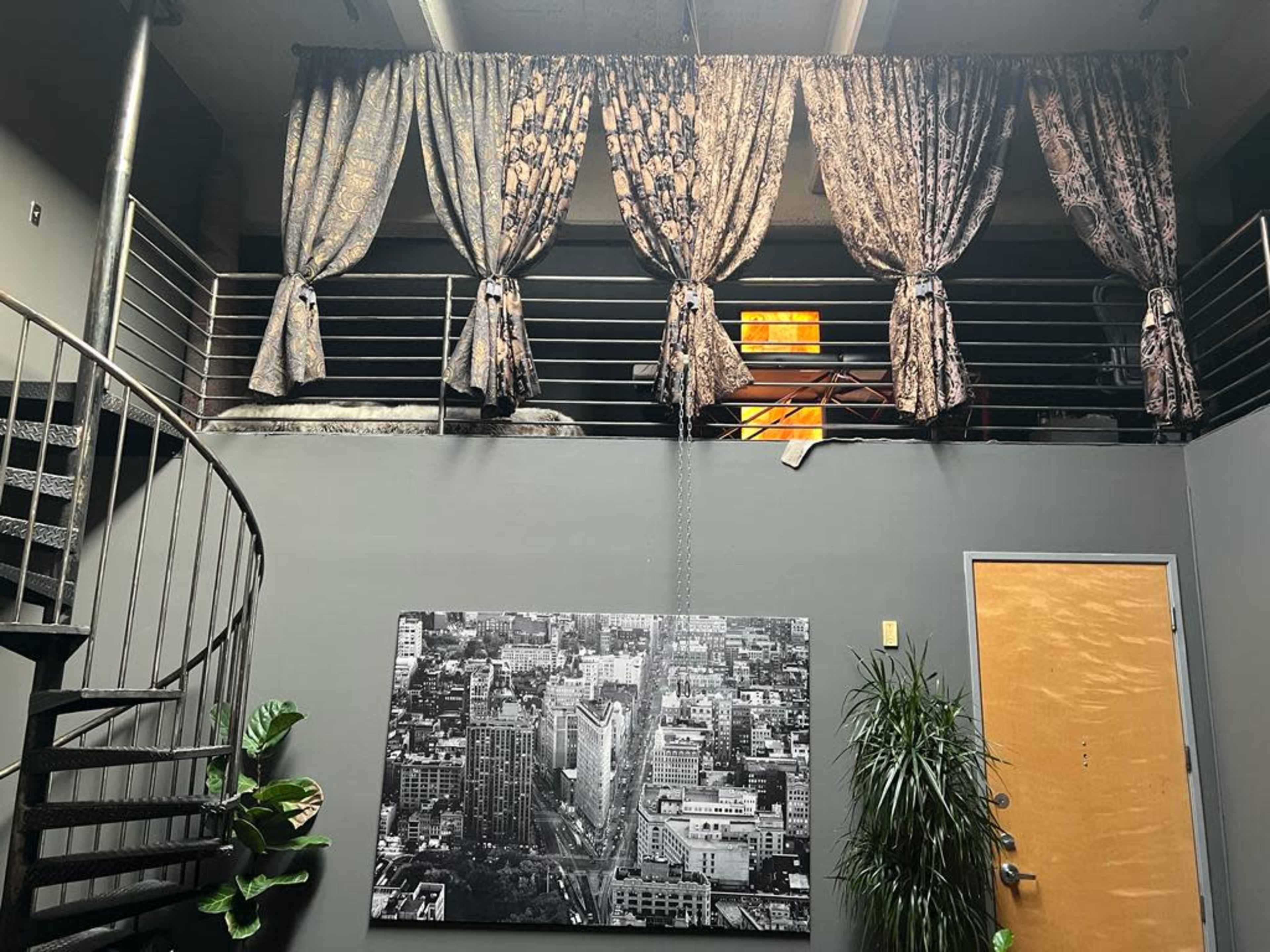 A modern interior space with a spiral staircase, decorative curtains draped over a upper-level railing, a large black-and-white cityscape photo on the wall, and potted plants in the foreground.