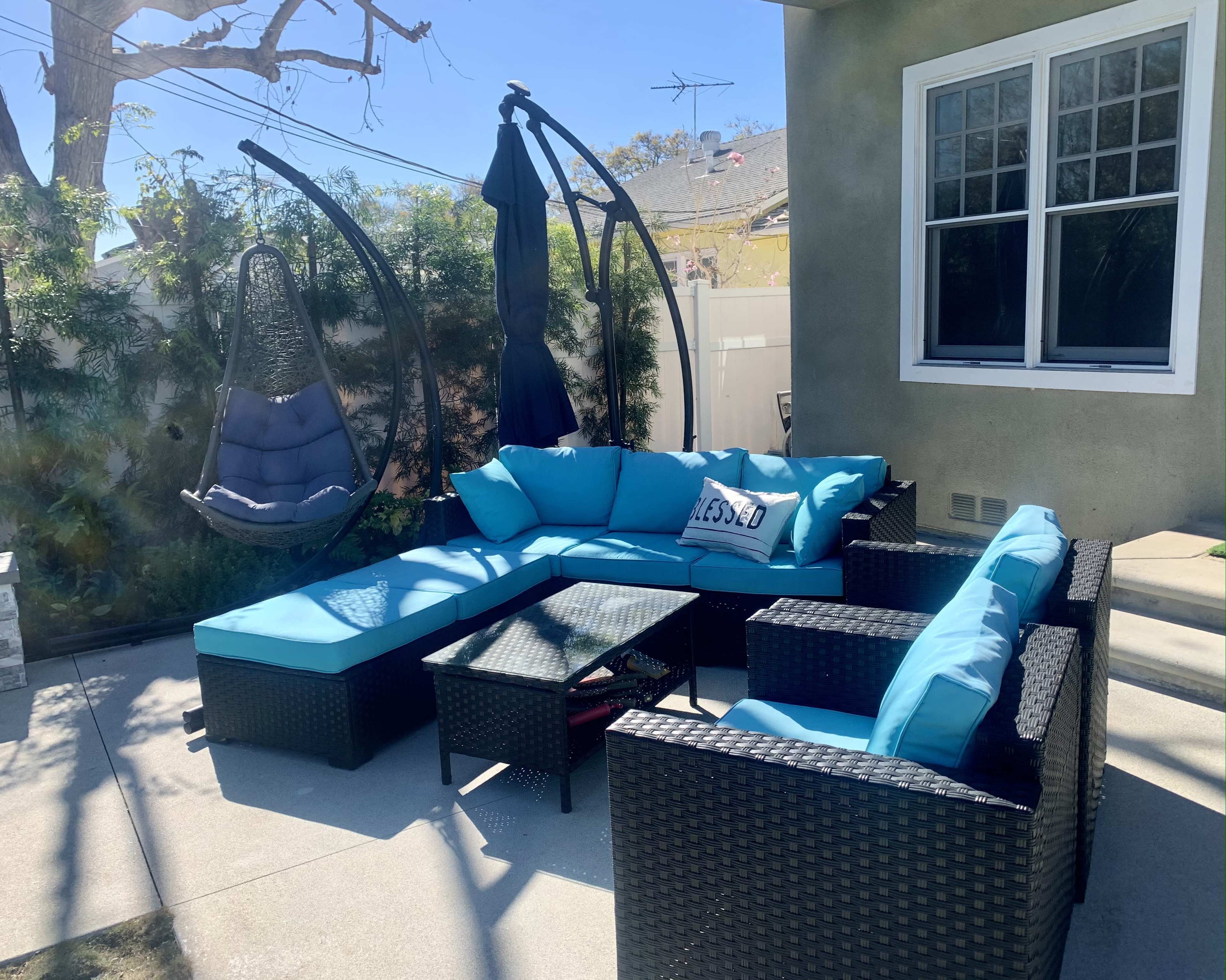 A patio area featuring a sectional sofa with bright blue cushions, a glass-top coffee table, and a hanging chair, set against a wall and surrounded by greenery.