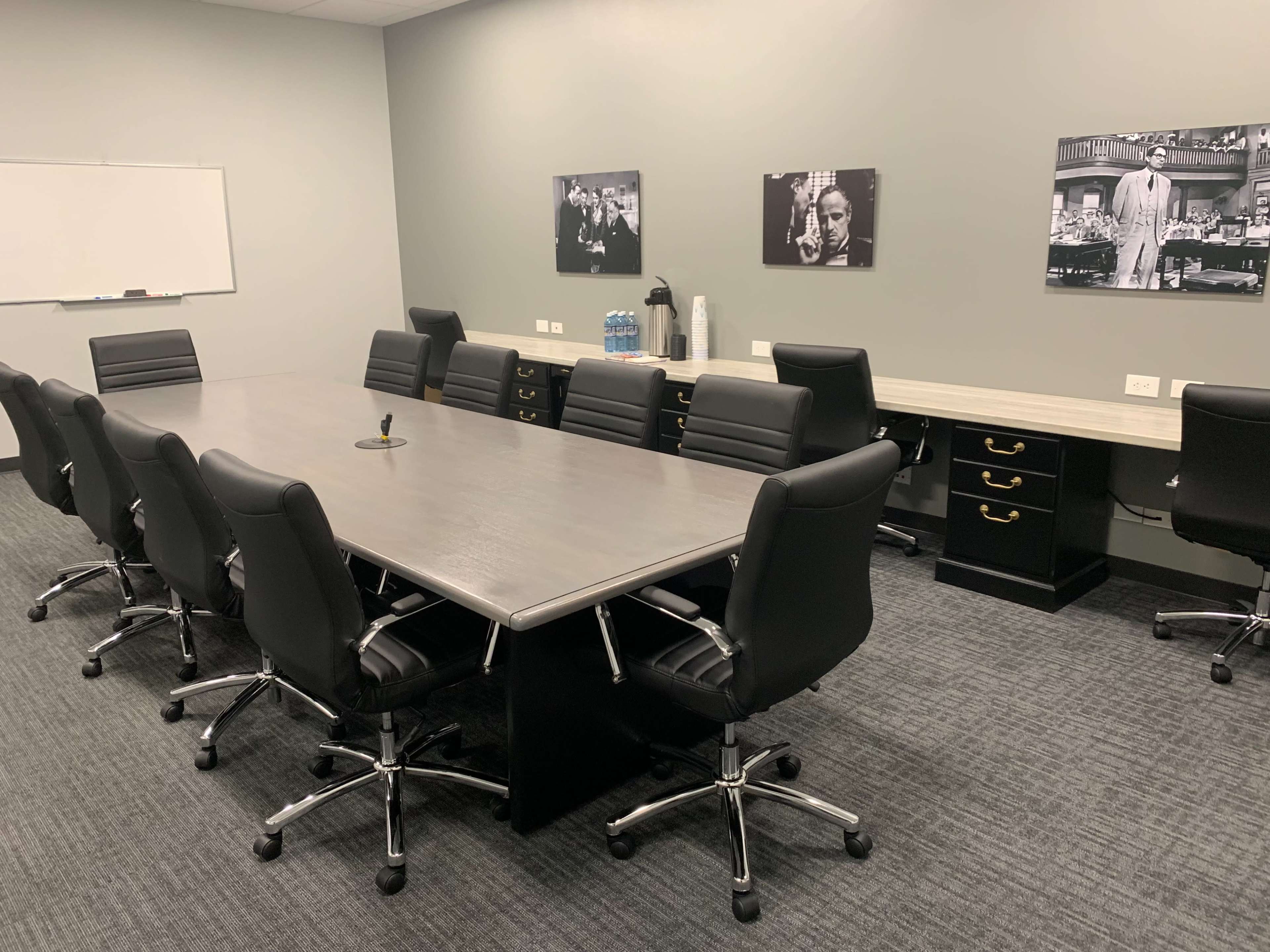 A conference room features a large rectangular table surrounded by black rolling chairs, with images on the walls and a counter along one side.