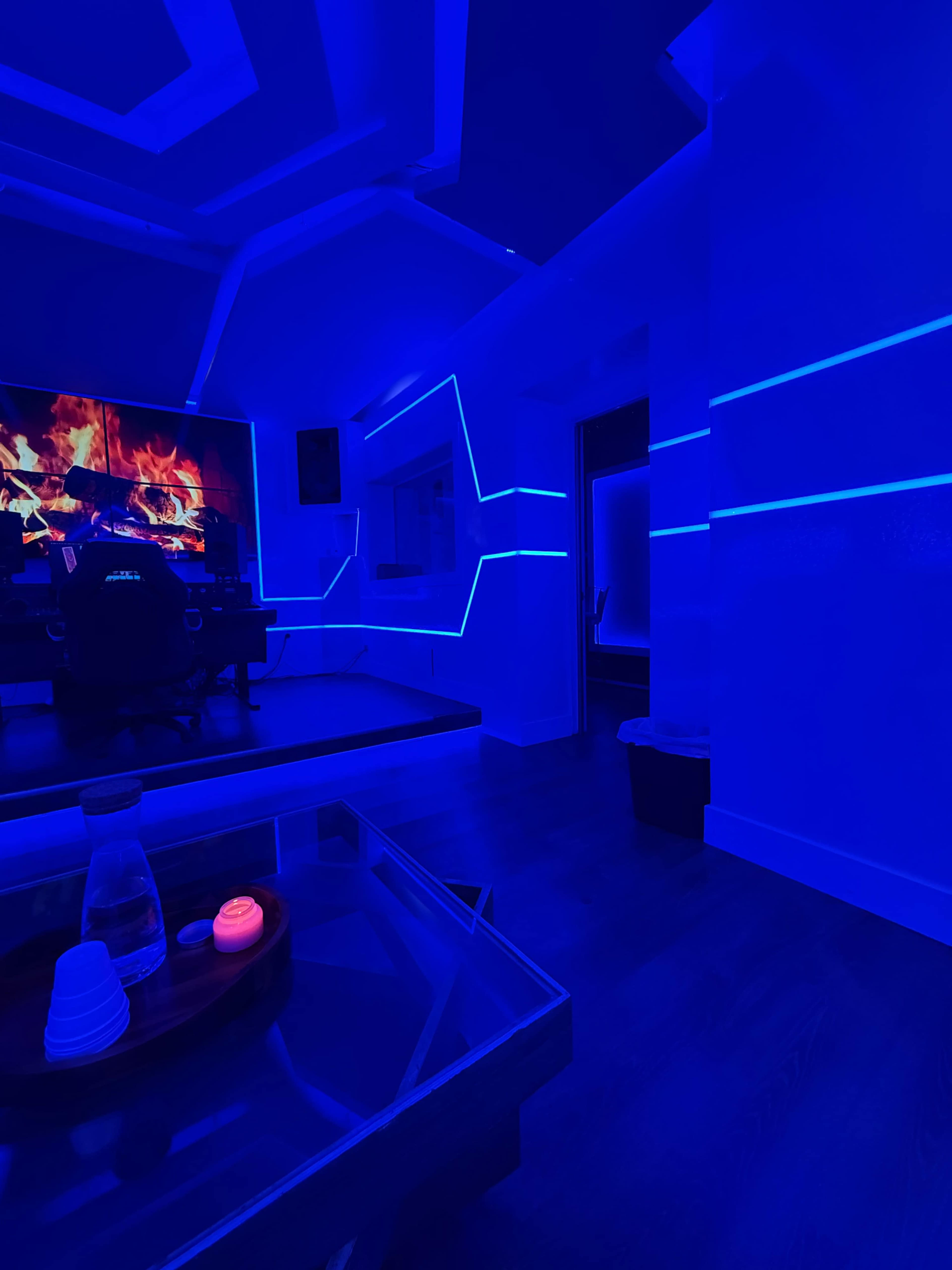 A modern interior space features blue LED lighting along the walls, a large screen displaying a fire image, and a glass coffee table with decorative items.