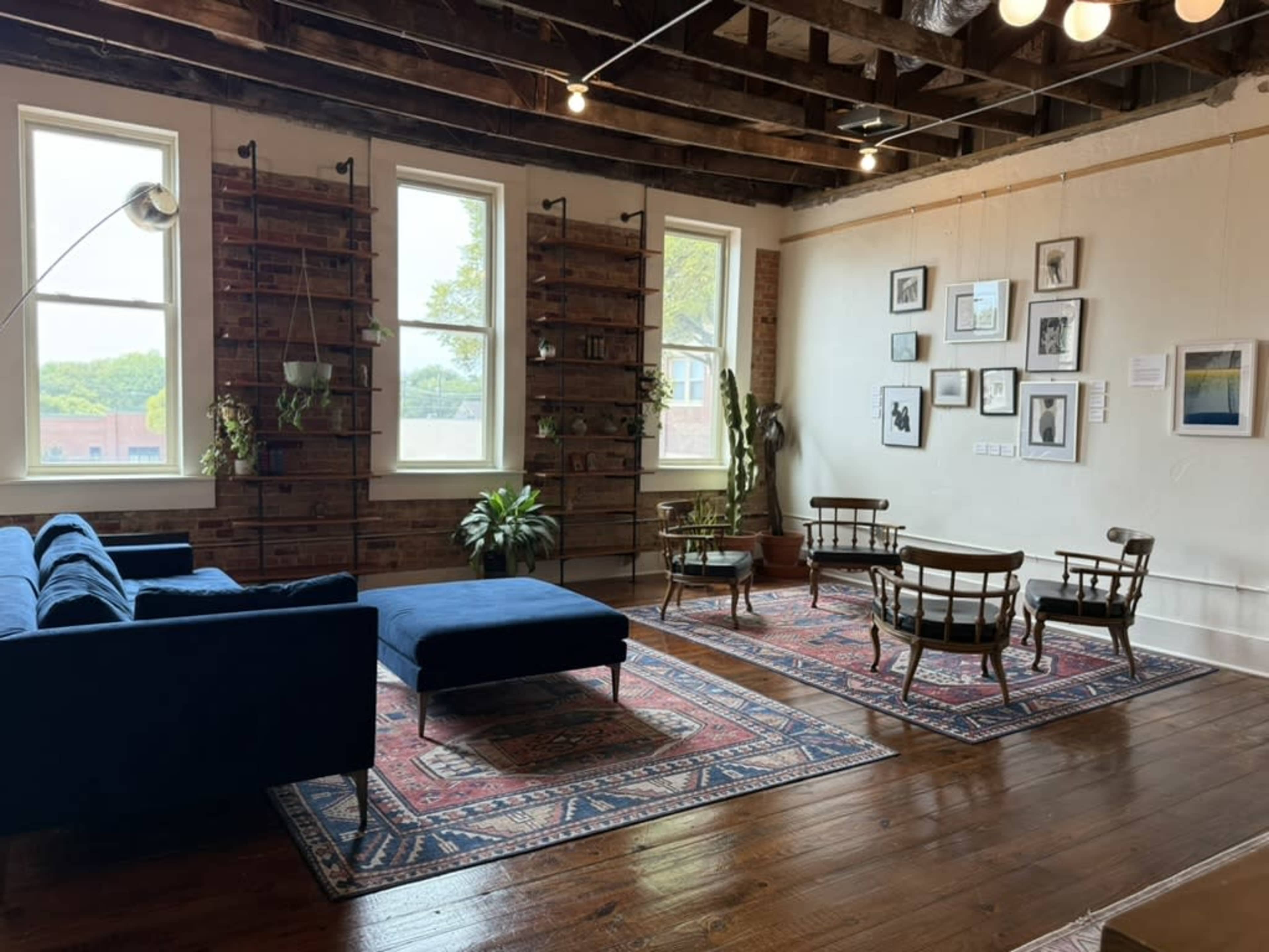 The image shows a spacious living area with a blue couch, armchairs, and a low ottoman, complemented by wooden shelves, plants, and framed pictures on the walls.