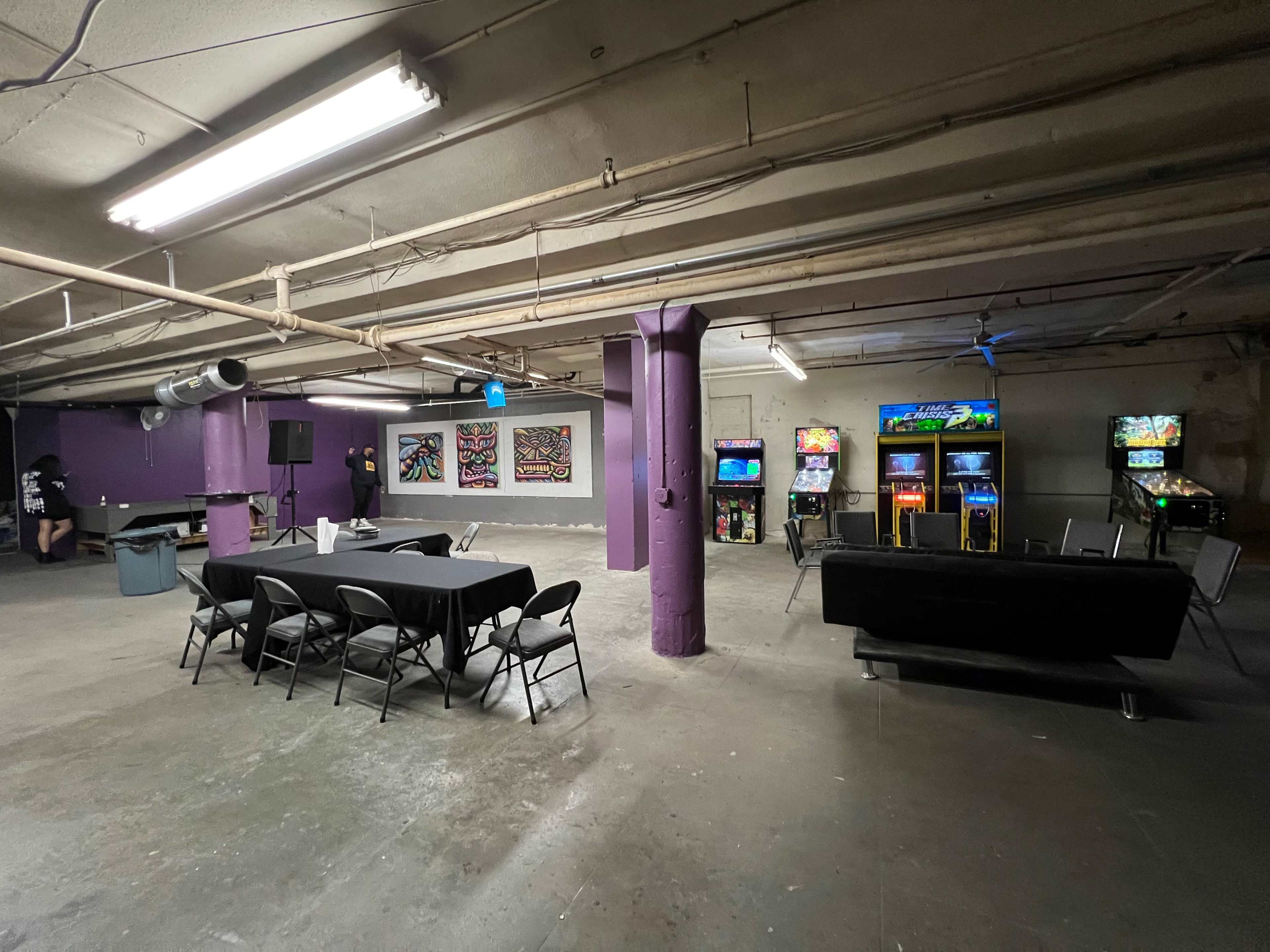 A dimly lit basement area with purple walls, featuring a table with black tablecloth and several chairs on one side, while arcade machines are arranged along the opposite wall.