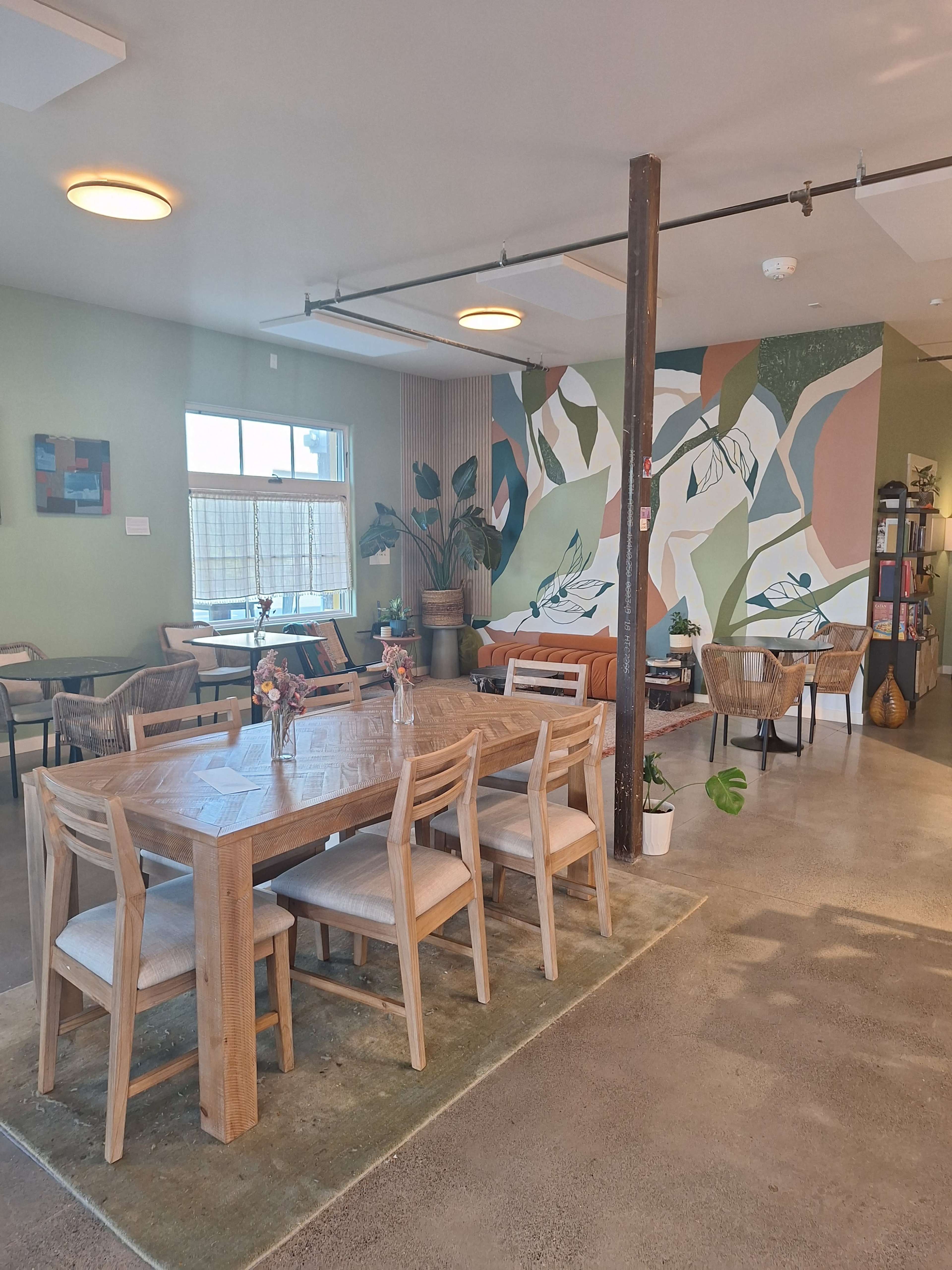 The image shows a modern café interior featuring a wooden dining table, chairs, a colorful mural on one wall, and various plants throughout the space.