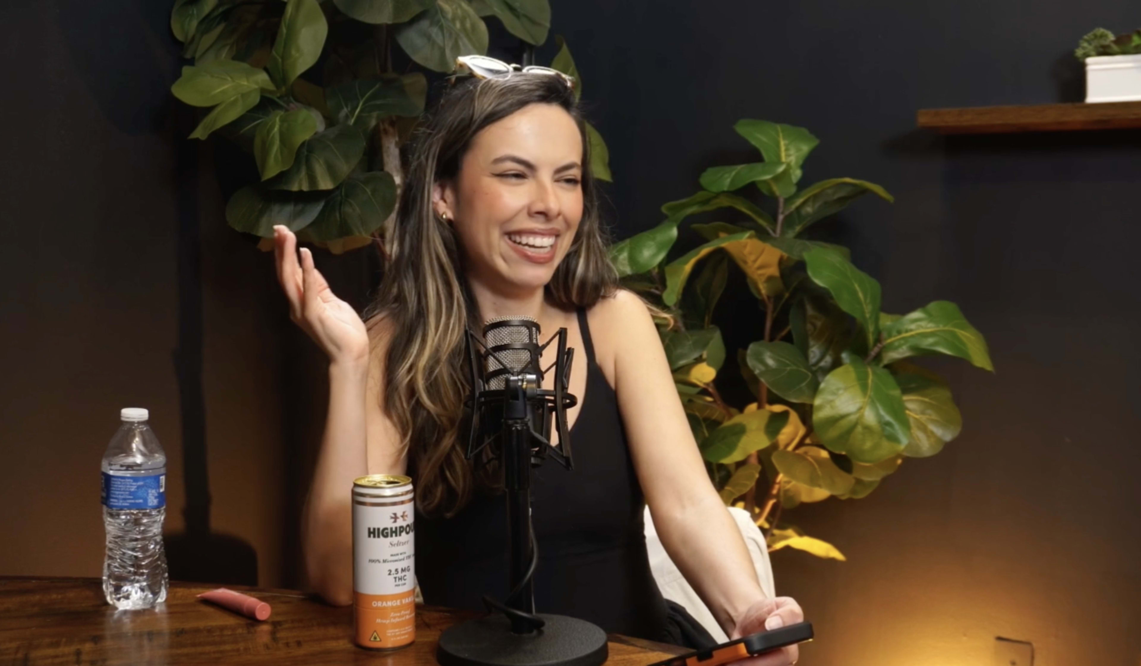 A woman sits at a table with a microphone, a water bottle, and a can of drink, surrounded by green plants.
