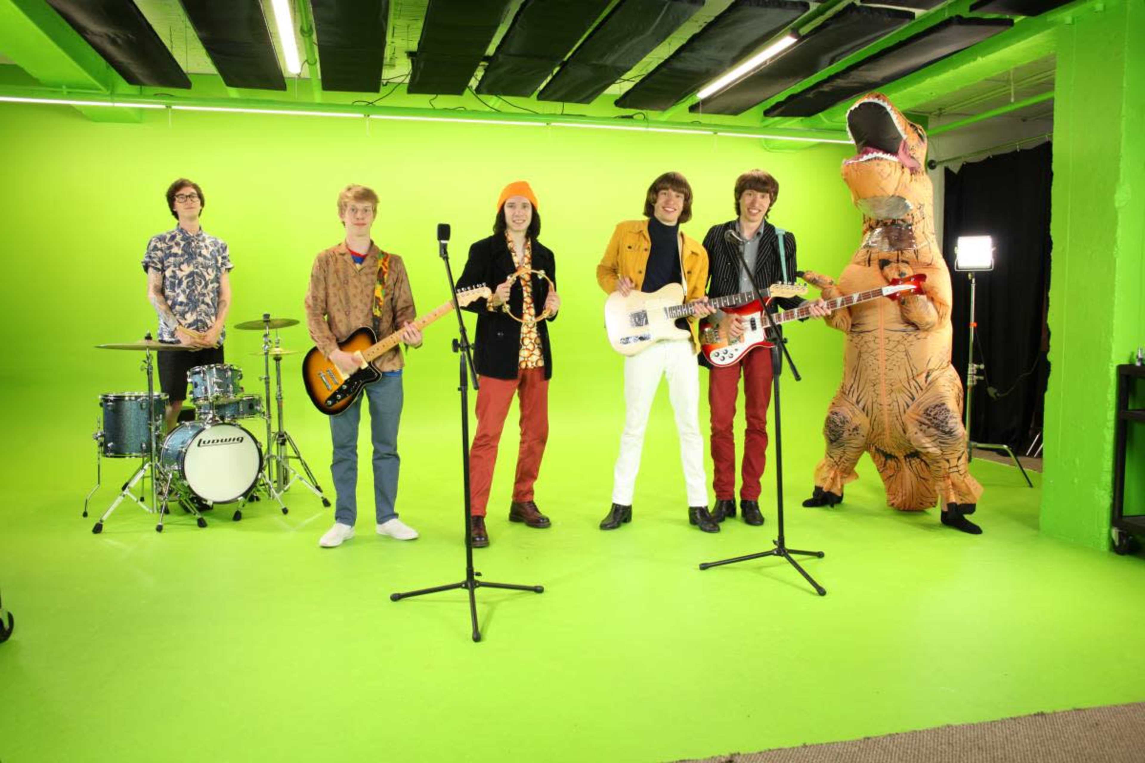 A six-member band stands on a bright green stage, with one member wearing a dinosaur costume, while others hold various musical instruments.