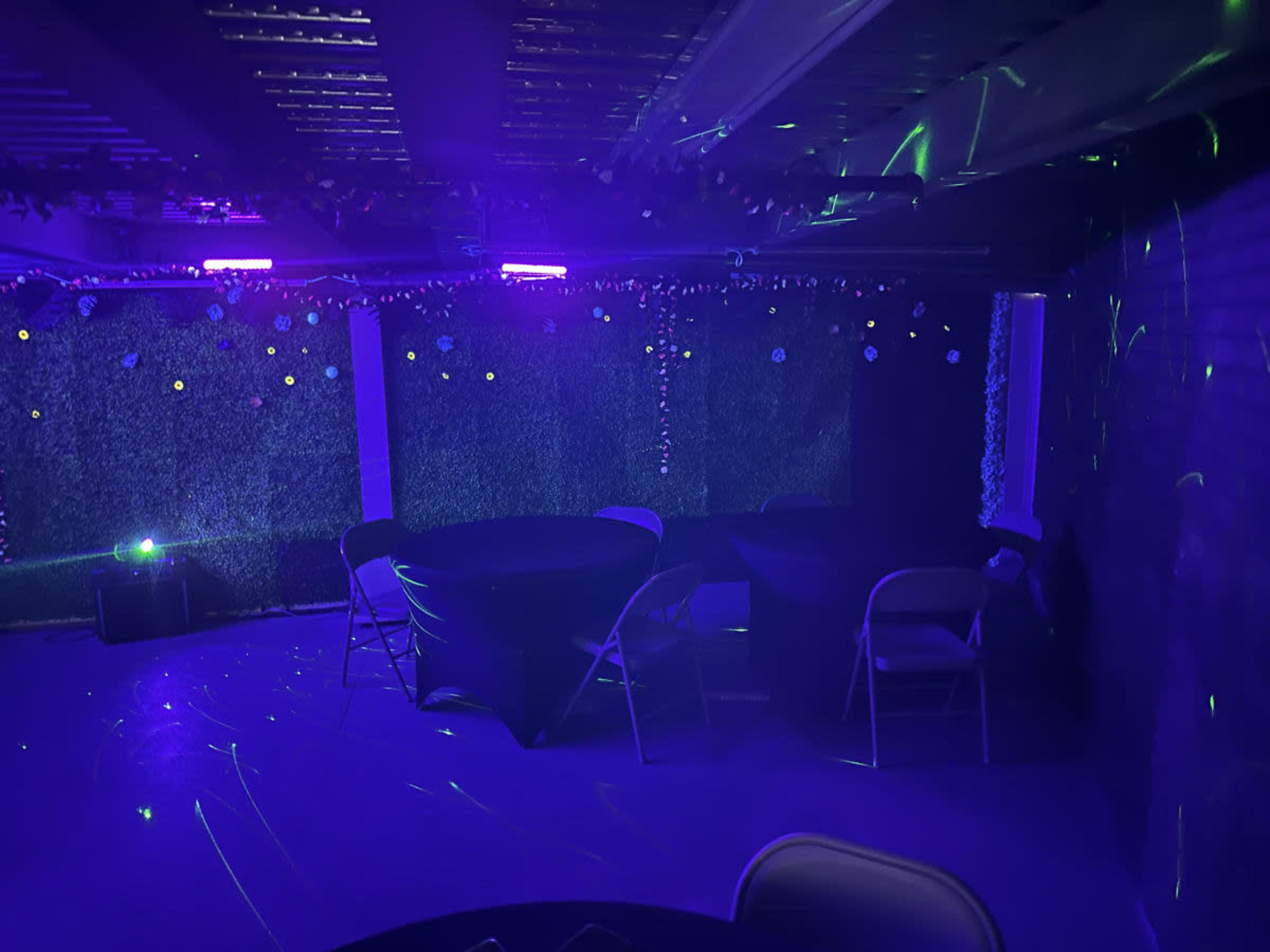 The image shows a dimly lit event space with purple and green lighting, featuring tables covered in black cloth and decorations along the walls.