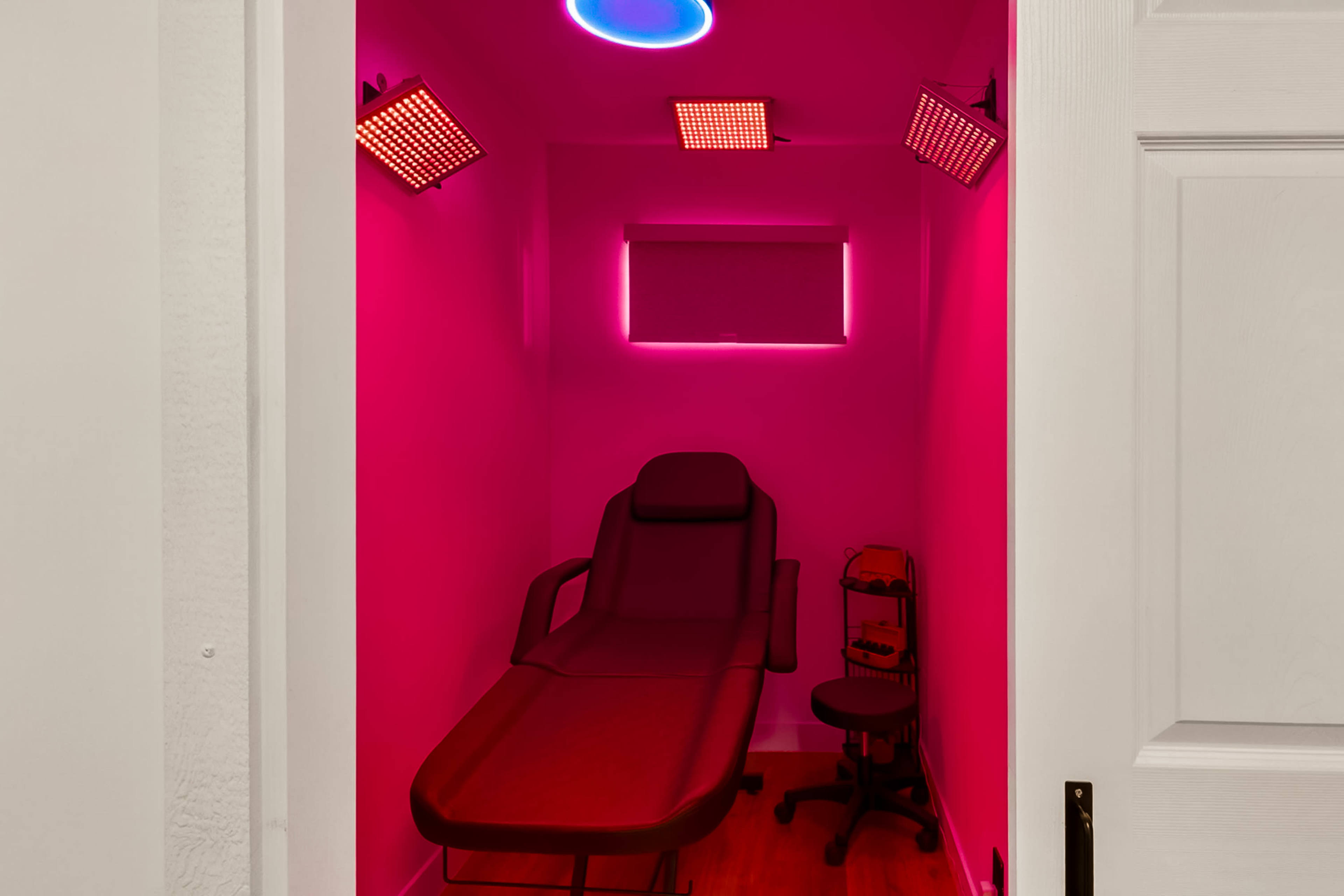 The image shows a small treatment room with a red-tinted interior, featuring a reclining chair and two light fixtures mounted on the walls.