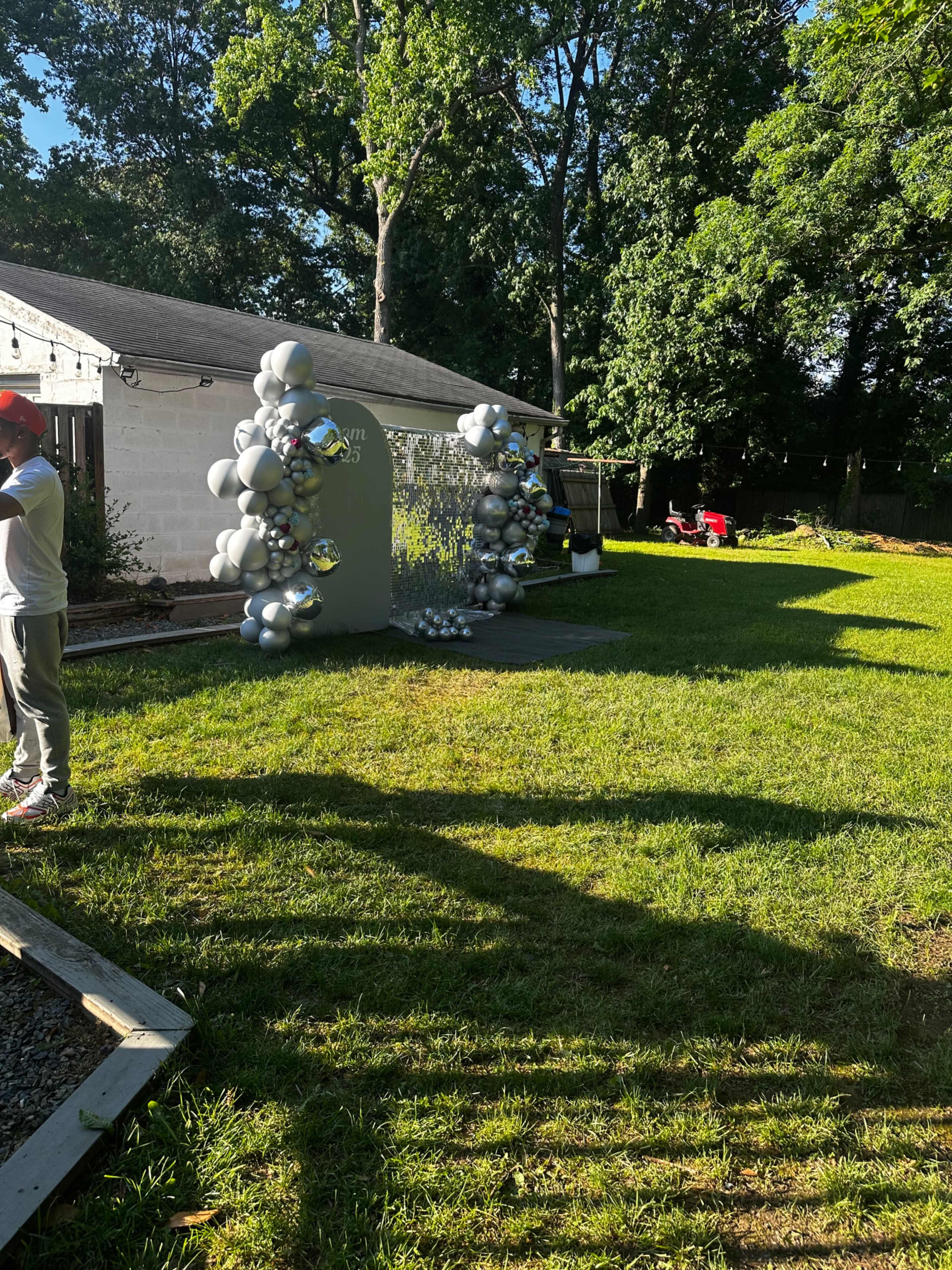 A backyard setting features a decorative balloon arrangement near a backdrop, with trees in the background and a few people engaged in activities.