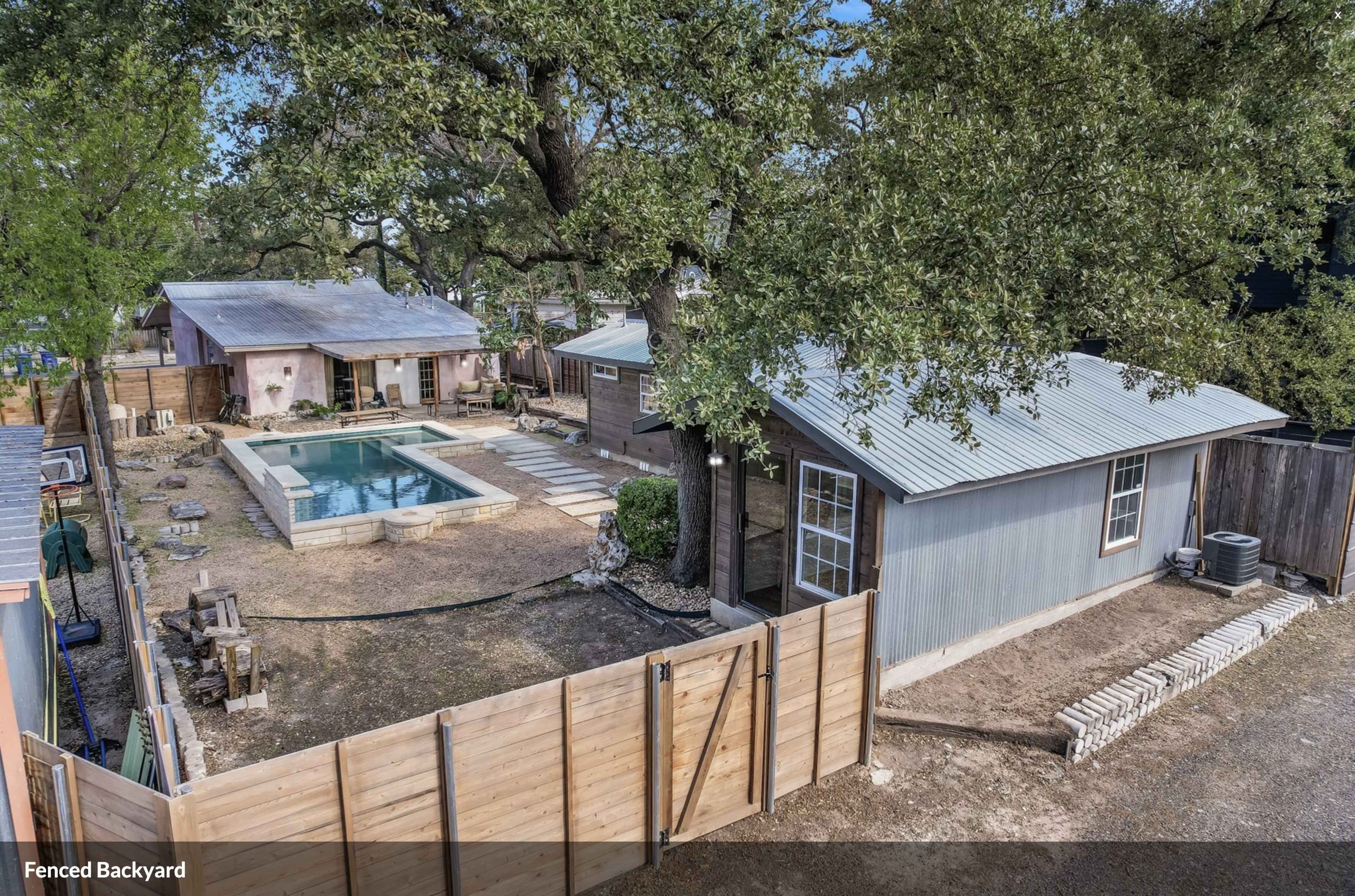 The image shows a fenced backyard featuring a swimming pool, a small house, a larger building in the background, and landscaped areas.