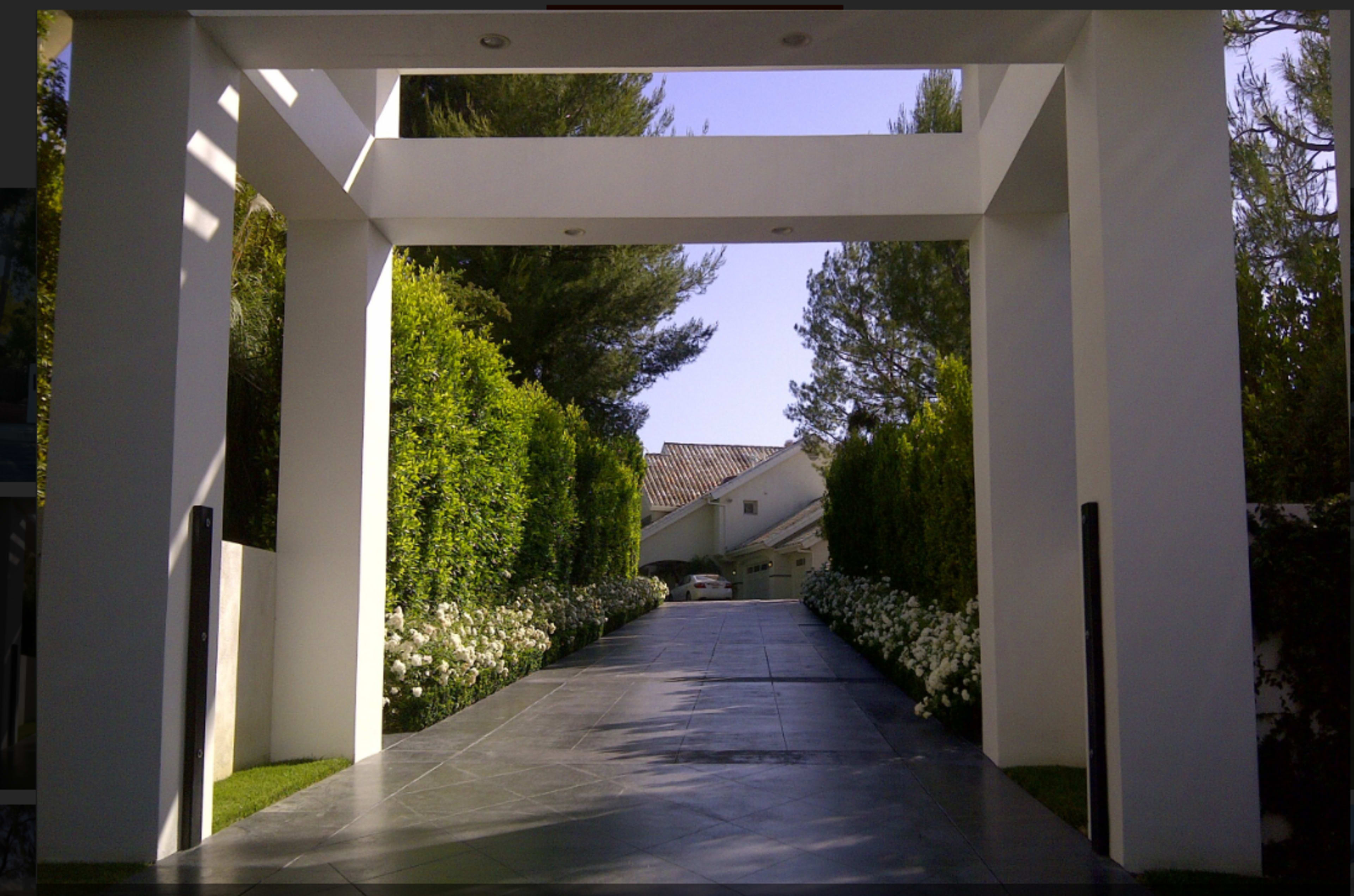 A modern driveway leads through a white archway, flanked by manicured hedges and flowering plants, toward a residential home in the background.