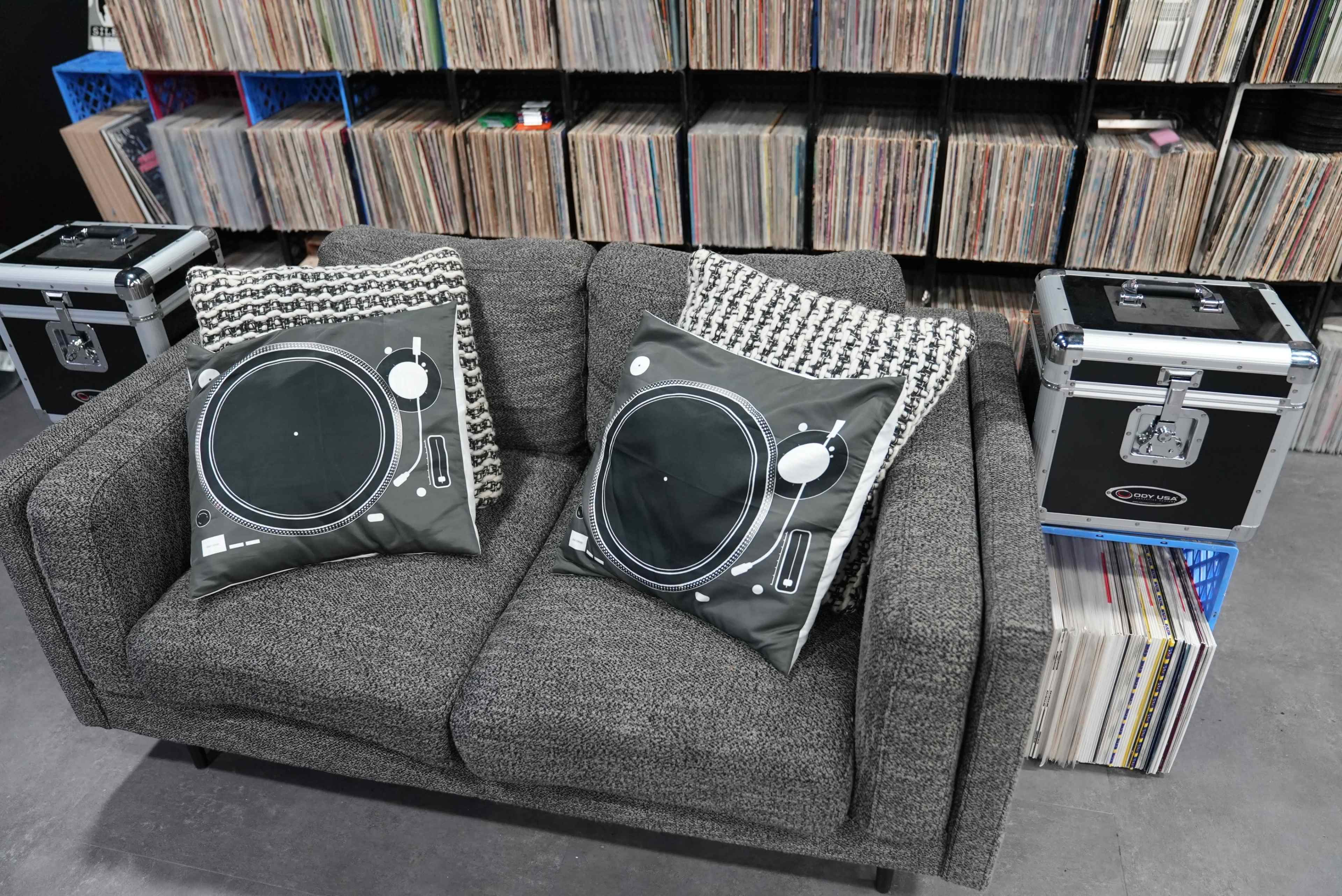 A gray couch with turntable-themed pillows is placed in front of a collection of vinyl records, flanked by two speaker cases.