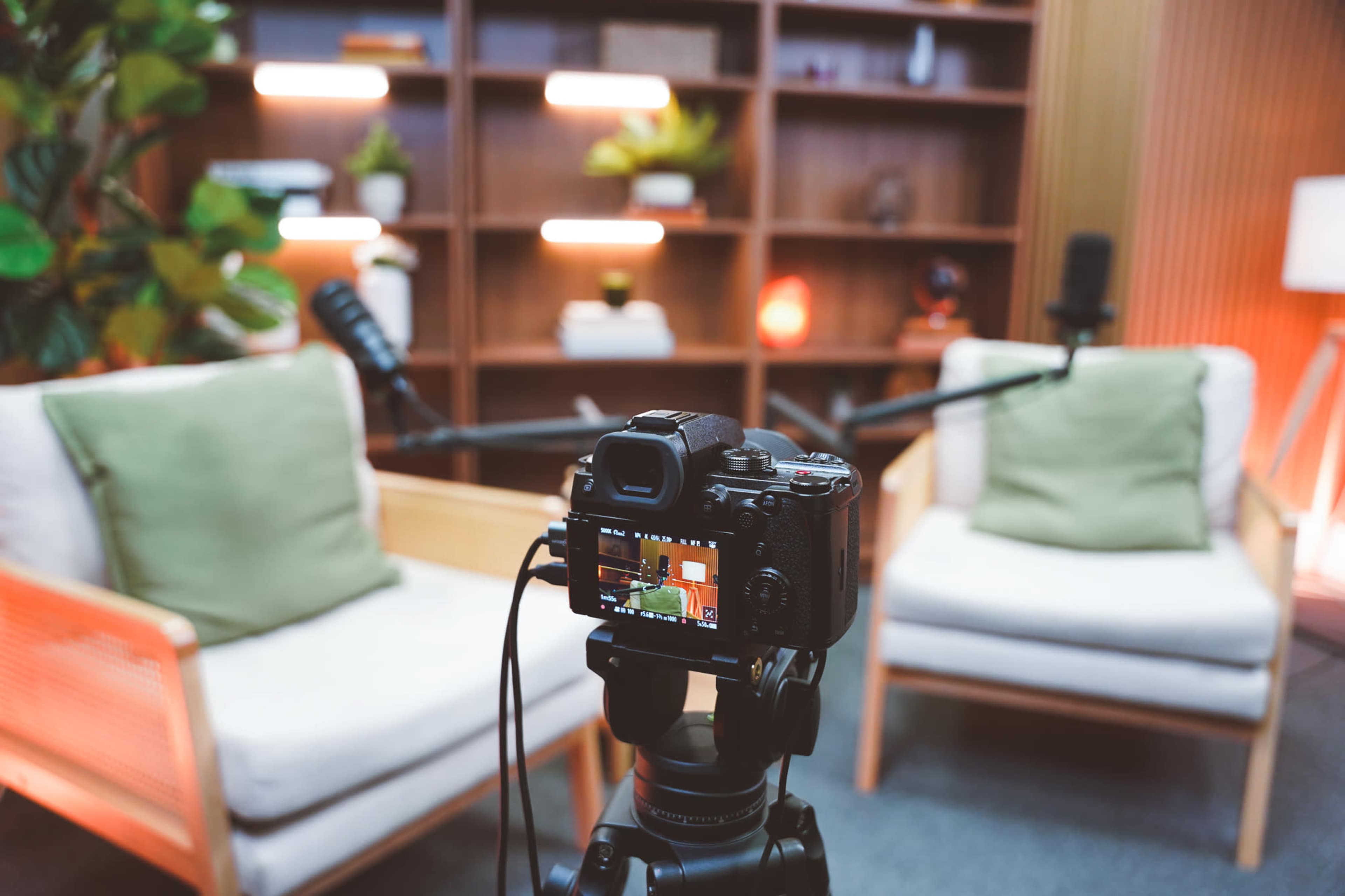 A camera on a tripod is focused on two empty chairs in a well-lit room with shelves containing plants and decorative items.