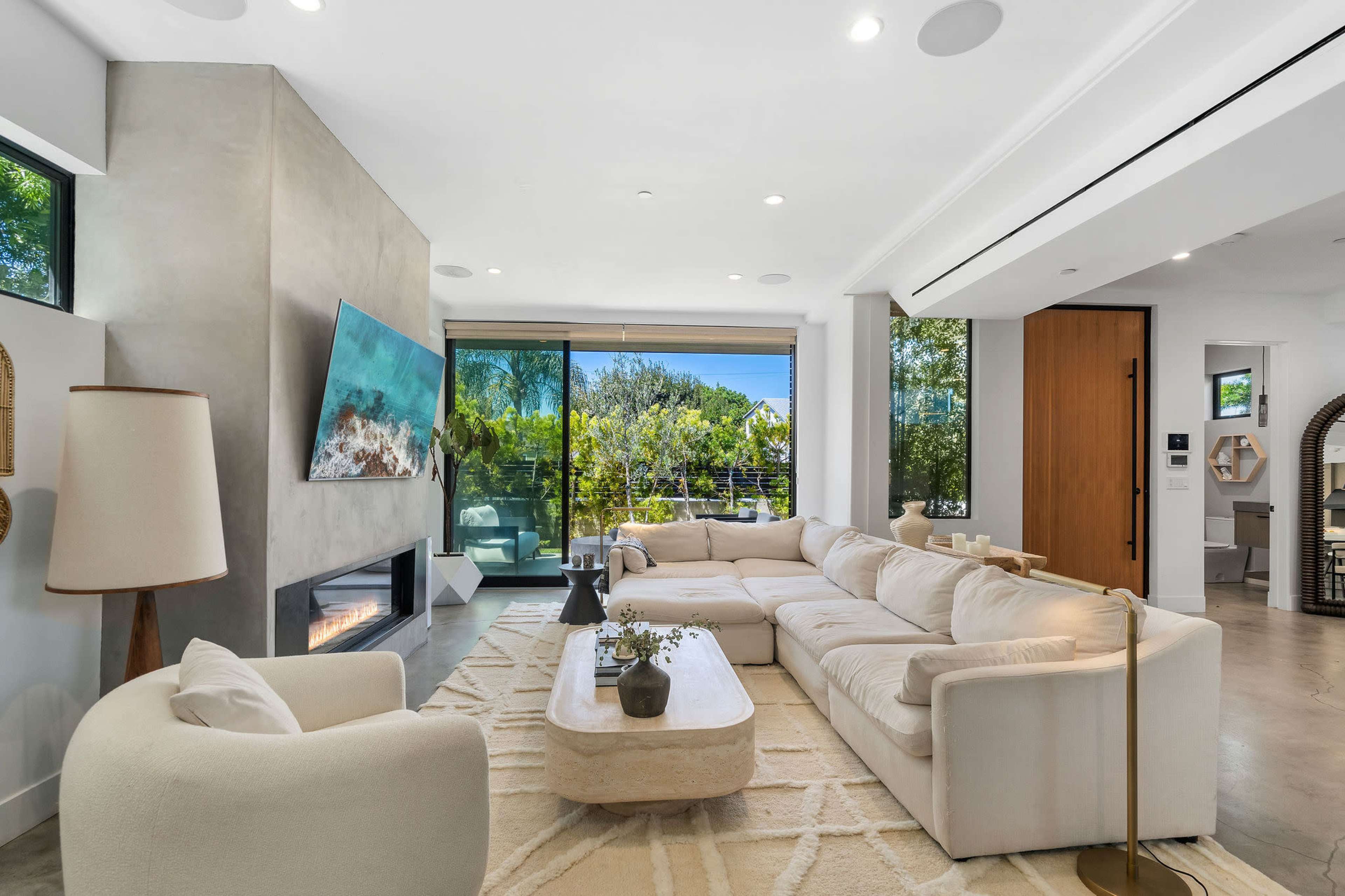 A modern living room with a spacious sectional sofa, a large television mounted on a concrete wall, and floor-to-ceiling windows allowing natural light to fill the space.