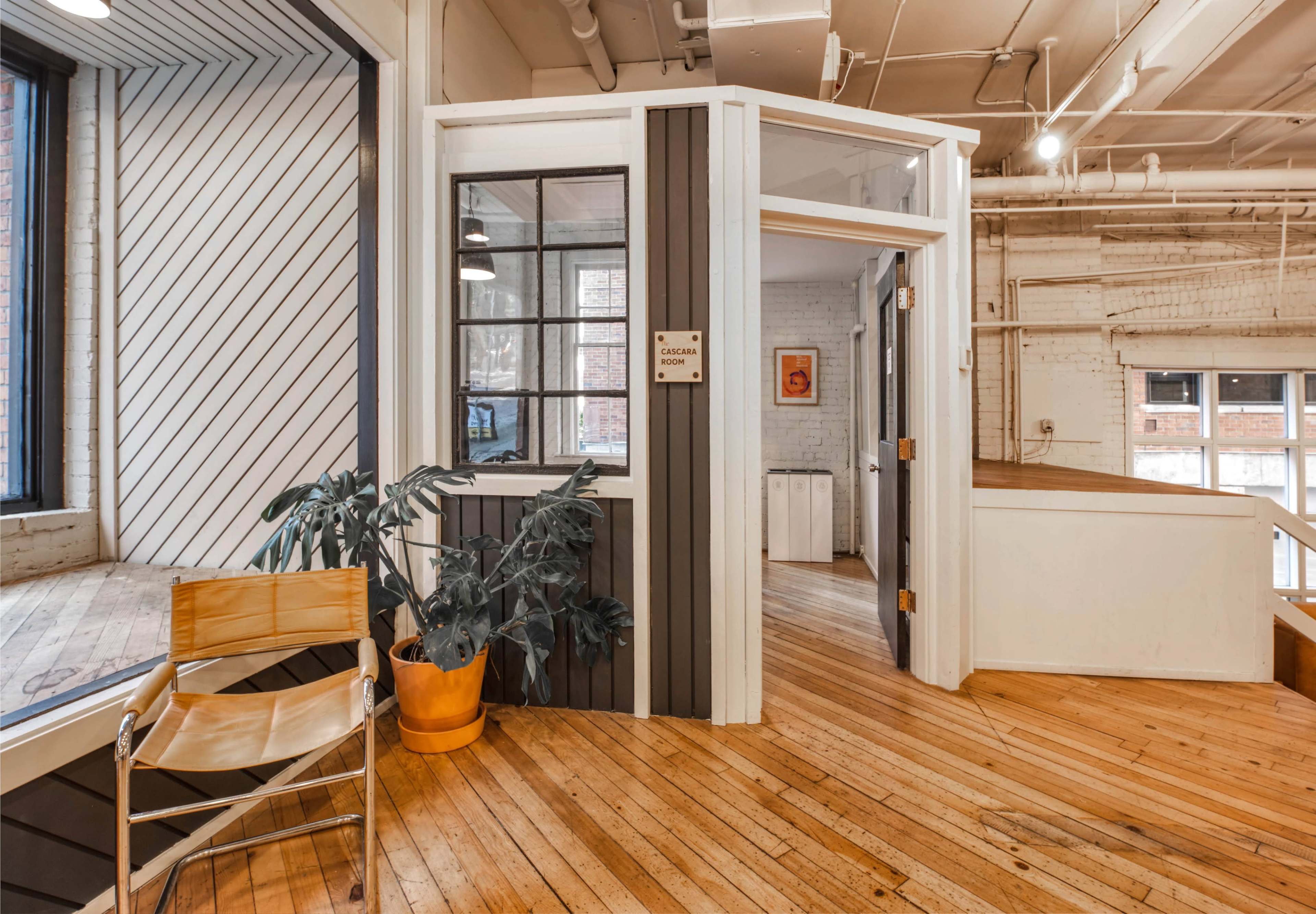 An interior space with wooden floors, divided by a glass-walled room, and a chair beside a potted plant.