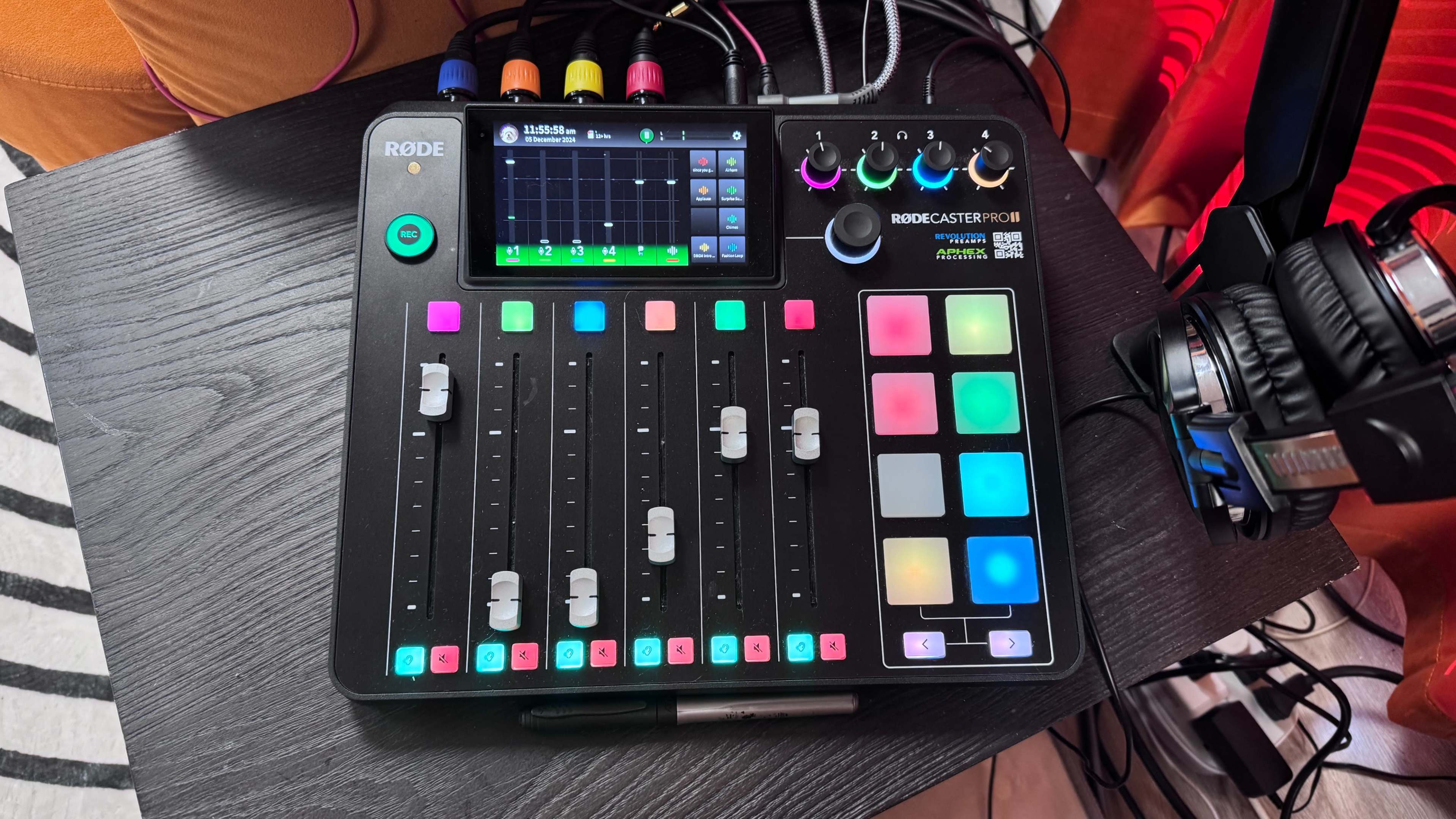 The image shows a Rodecaster Pro II audio mixing console with multiple sliders, buttons, and a digital display on a table.