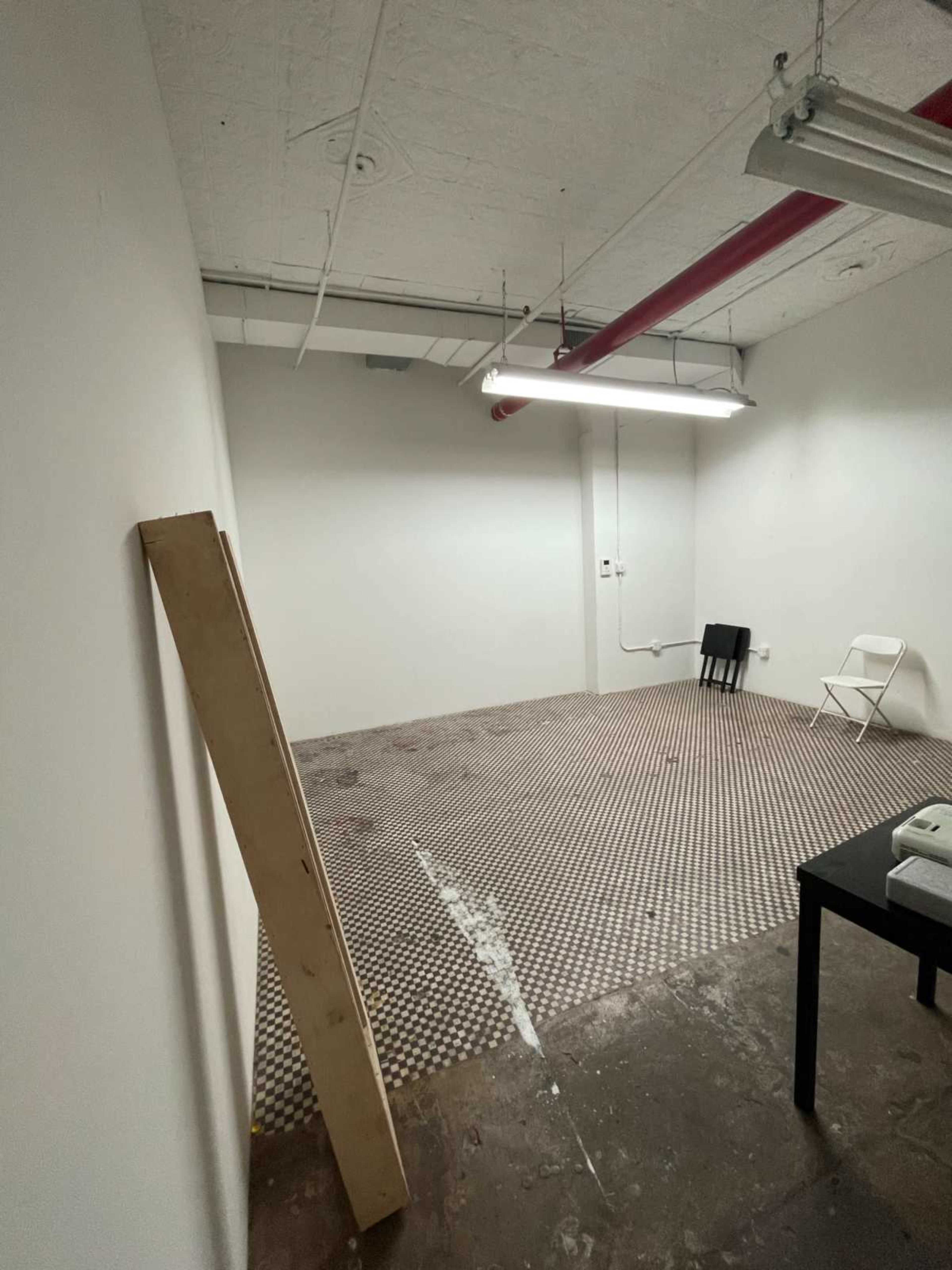 The image shows an empty room with a patterned floor, a chair in the corner, a table, and overhead lighting.