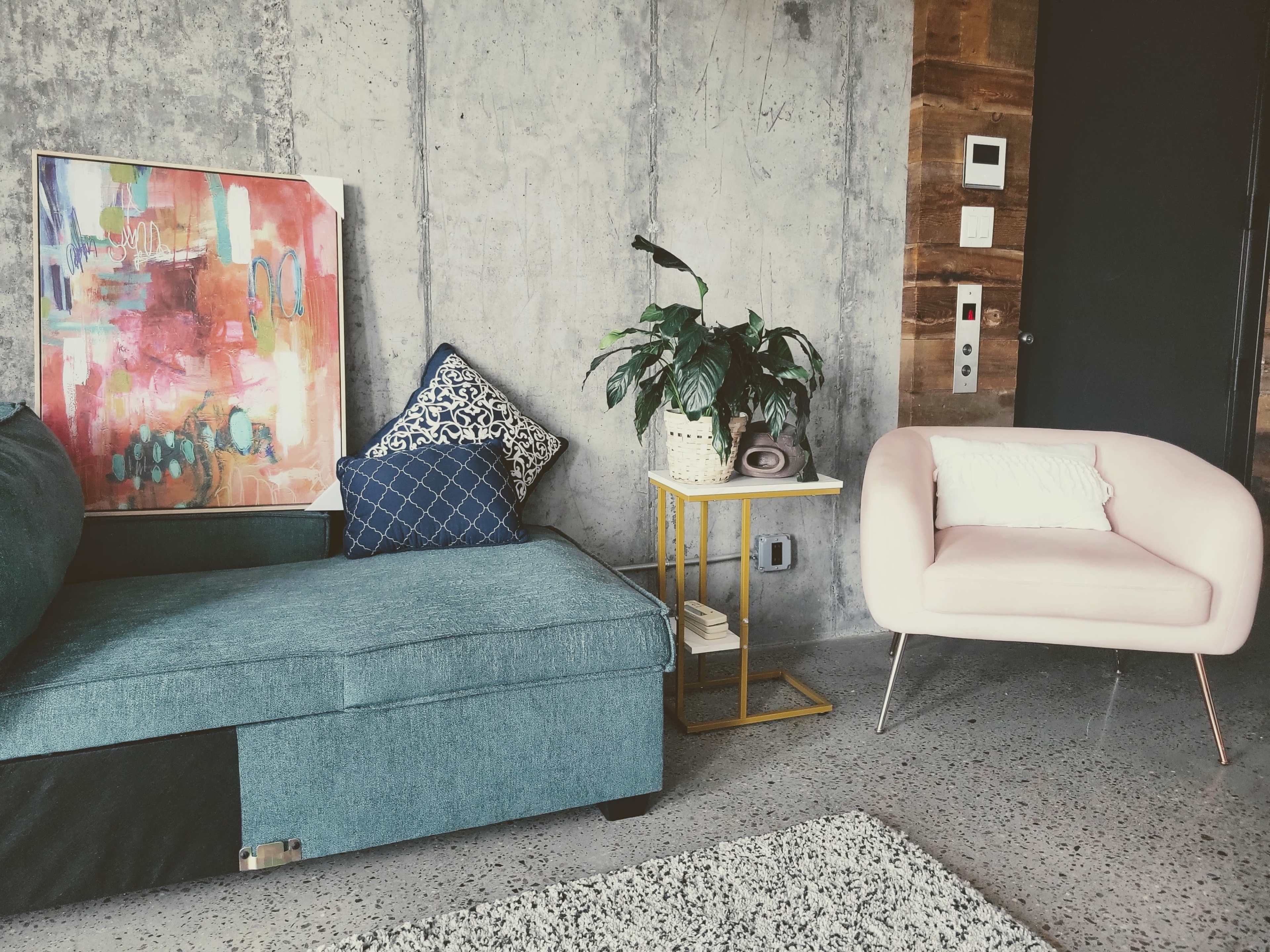 The scene features a teal sectional sofa, a pink upholstered armchair, a small side table, a potted plant, and a colorful abstract painting against a concrete wall.