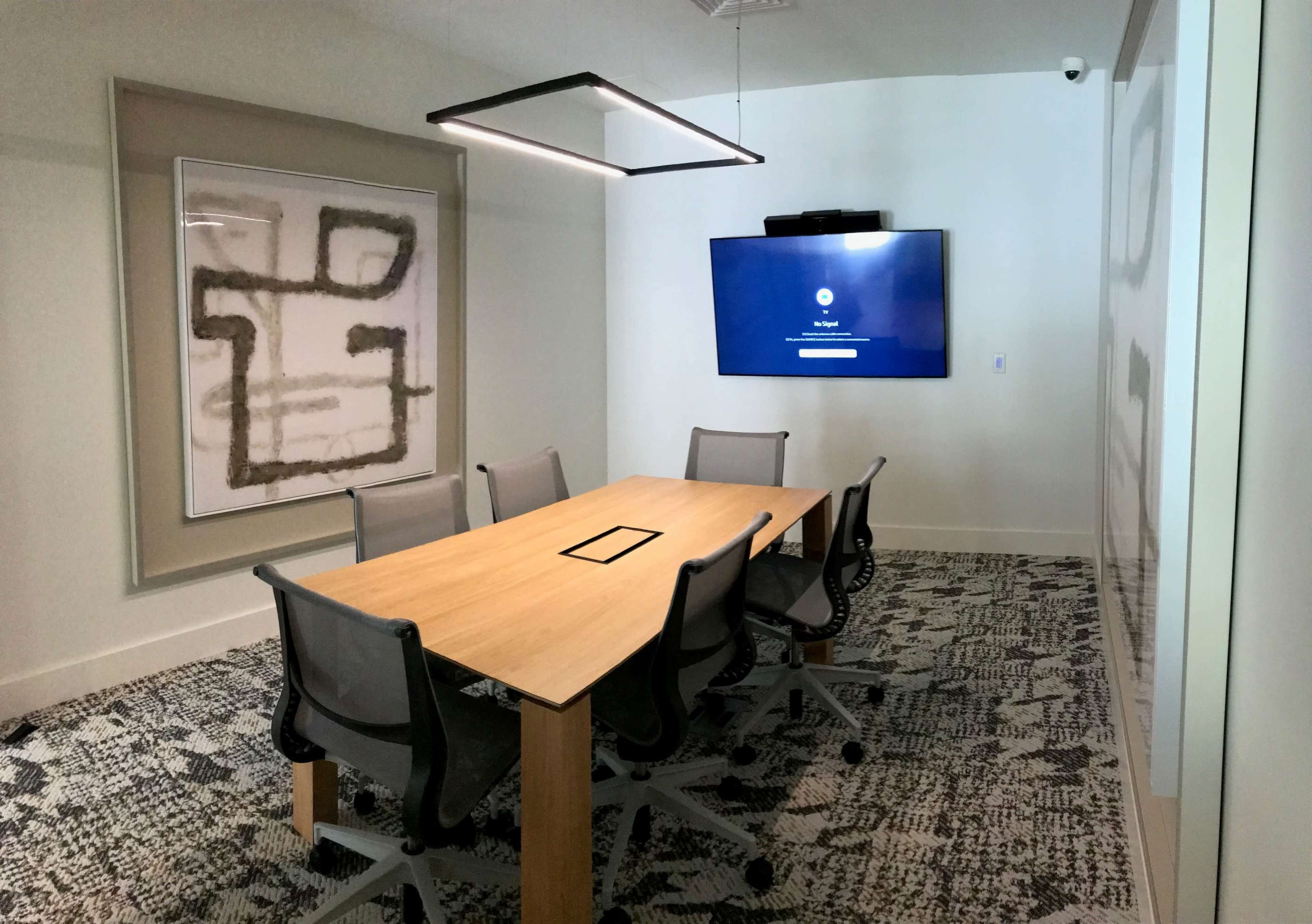 A modern conference room features a long wooden table surrounded by six ergonomic chairs, with a large flat-screen TV mounted on the wall displaying a login screen.