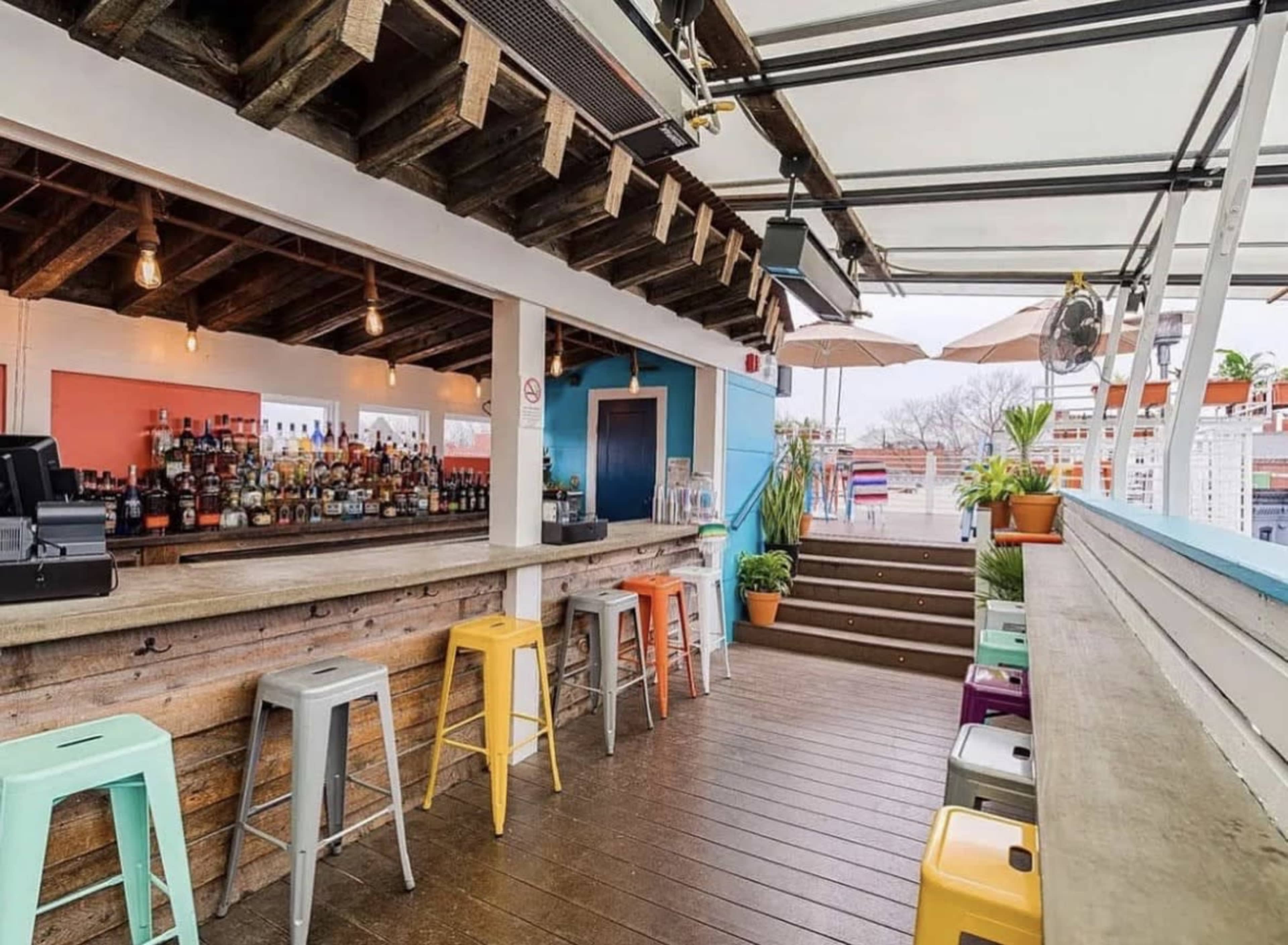 The image shows a bright bar area with a wooden countertop, colorful high stools, and a variety of bottled drinks displayed on shelves, set under a covered patio with an open space leading to stairs.