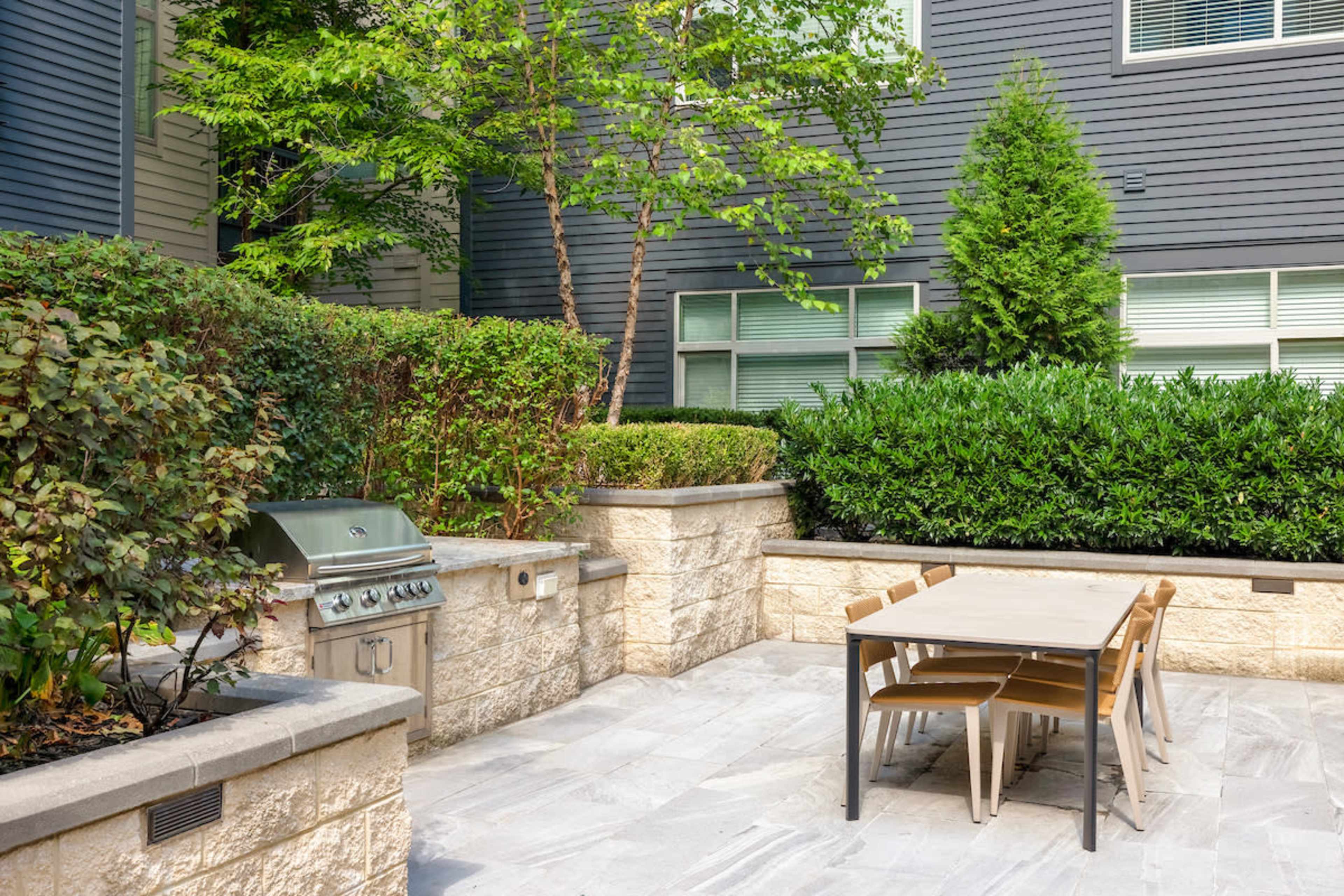 The image shows an outdoor patio area featuring a grill and a table surrounded by landscaped greenery and stone walls.