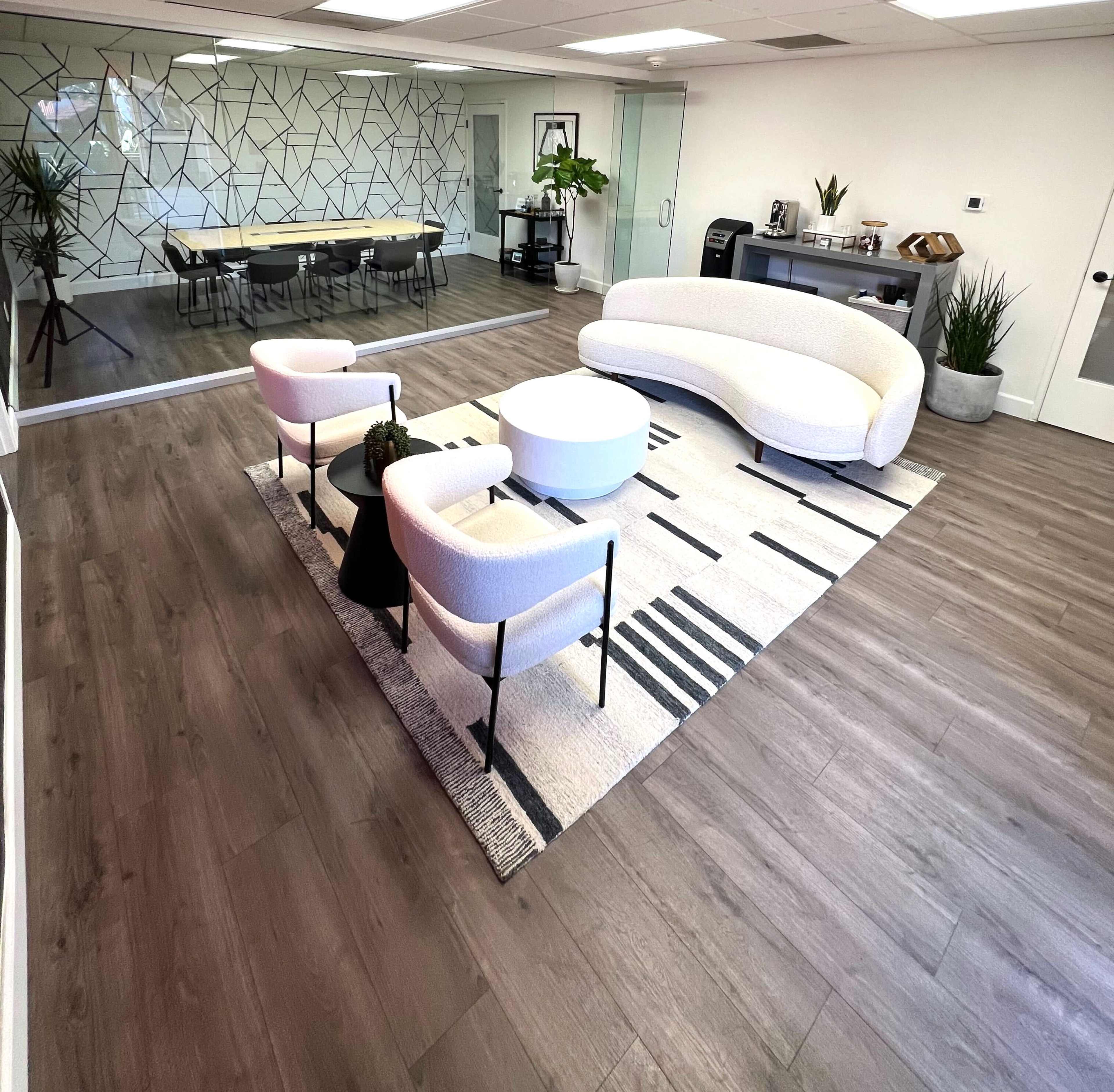 A modern office space features a curved white sofa, two accent chairs, and a round coffee table on a patterned rug, with a conference table in the background and greenery throughout.