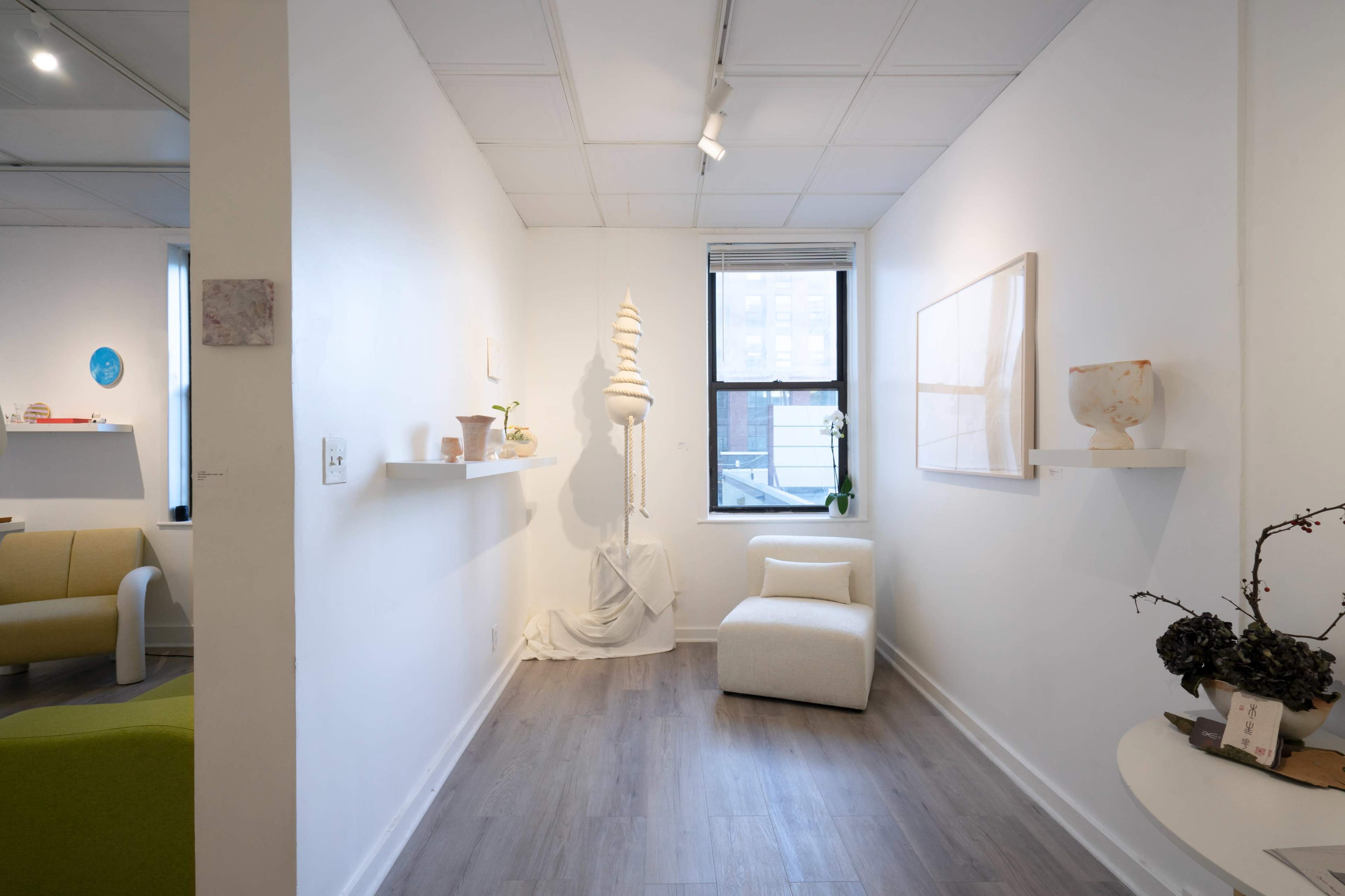 The image shows a contemporary art gallery interior featuring white walls, wooden flooring, a minimalist sculpture, and various artworks displayed on shelves.