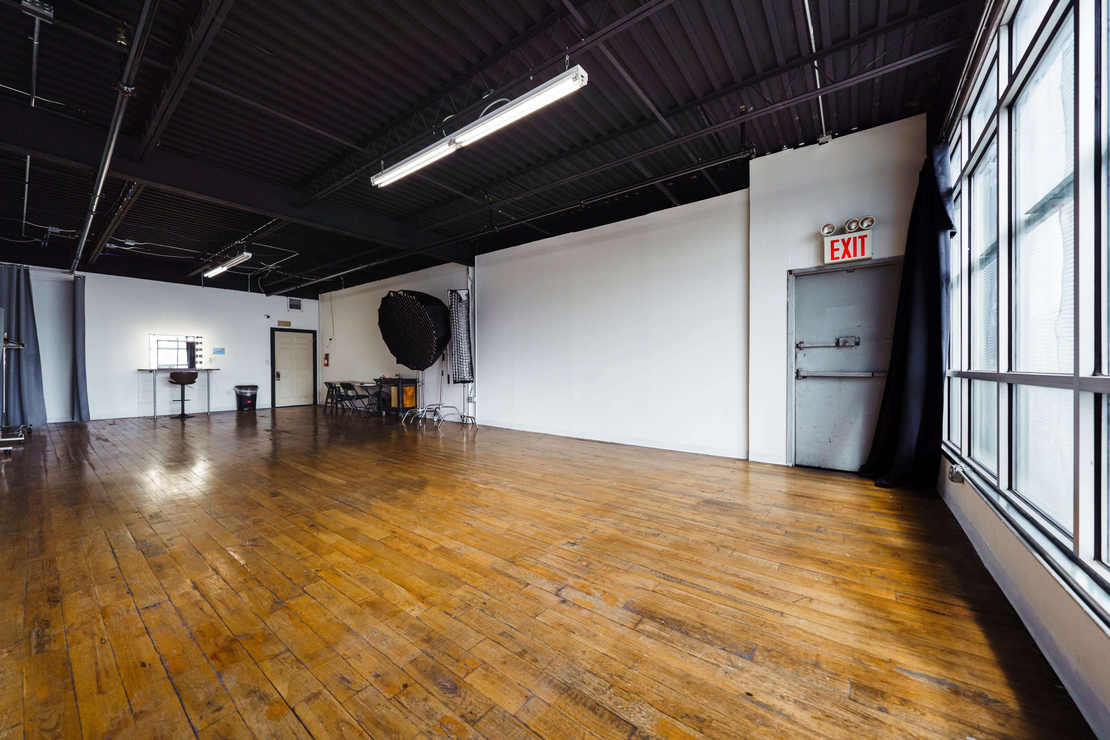 The image shows a spacious, empty studio with wooden floors, large windows, and various photography equipment set up in the corners.
