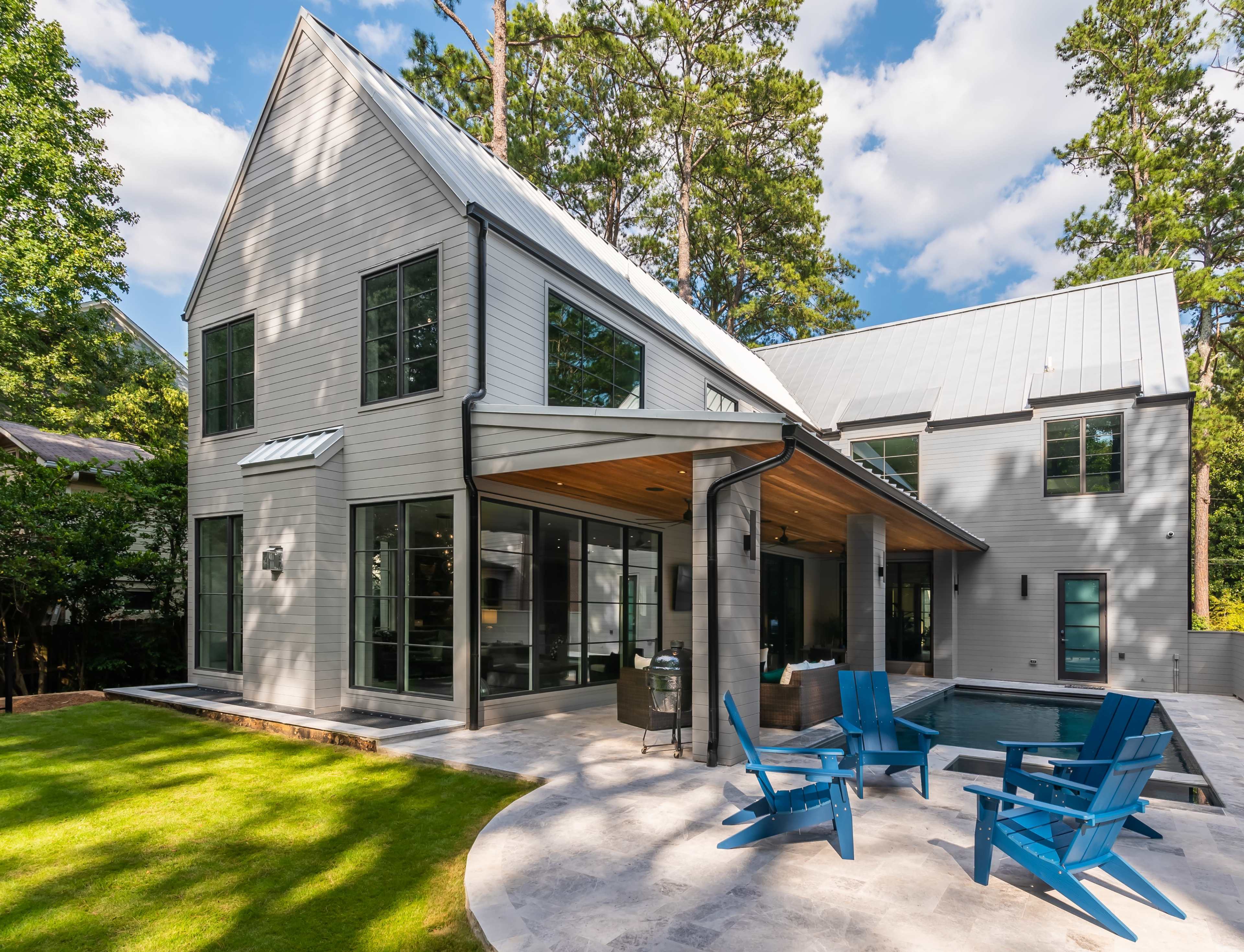 The image shows a contemporary house with a gray exterior, large windows, and a spacious patio area featuring blue Adirondack chairs beside a small pool.