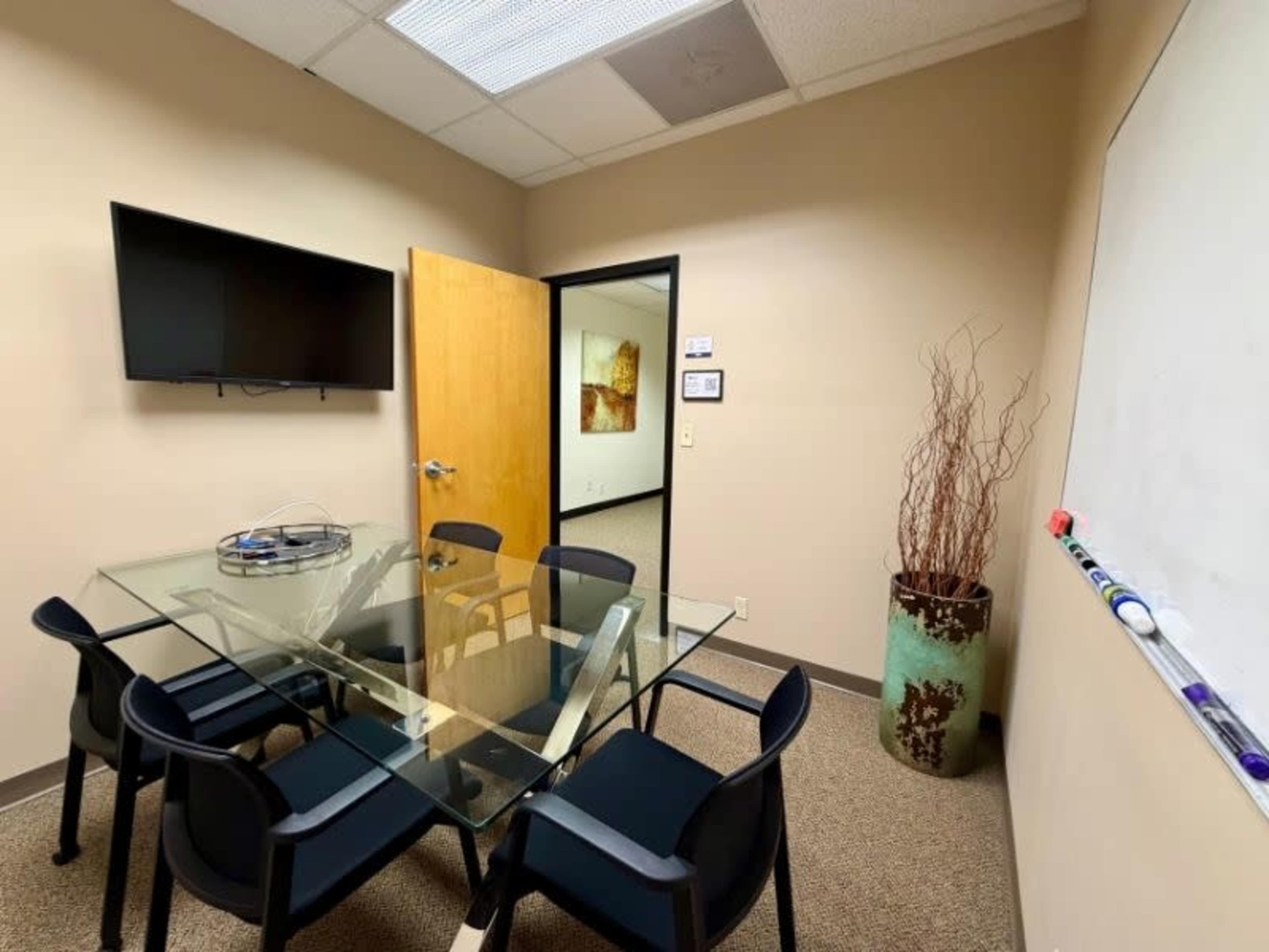 The image shows a small meeting room with a glass table surrounded by black chairs, a wall-mounted television, and a whiteboard on one side.
