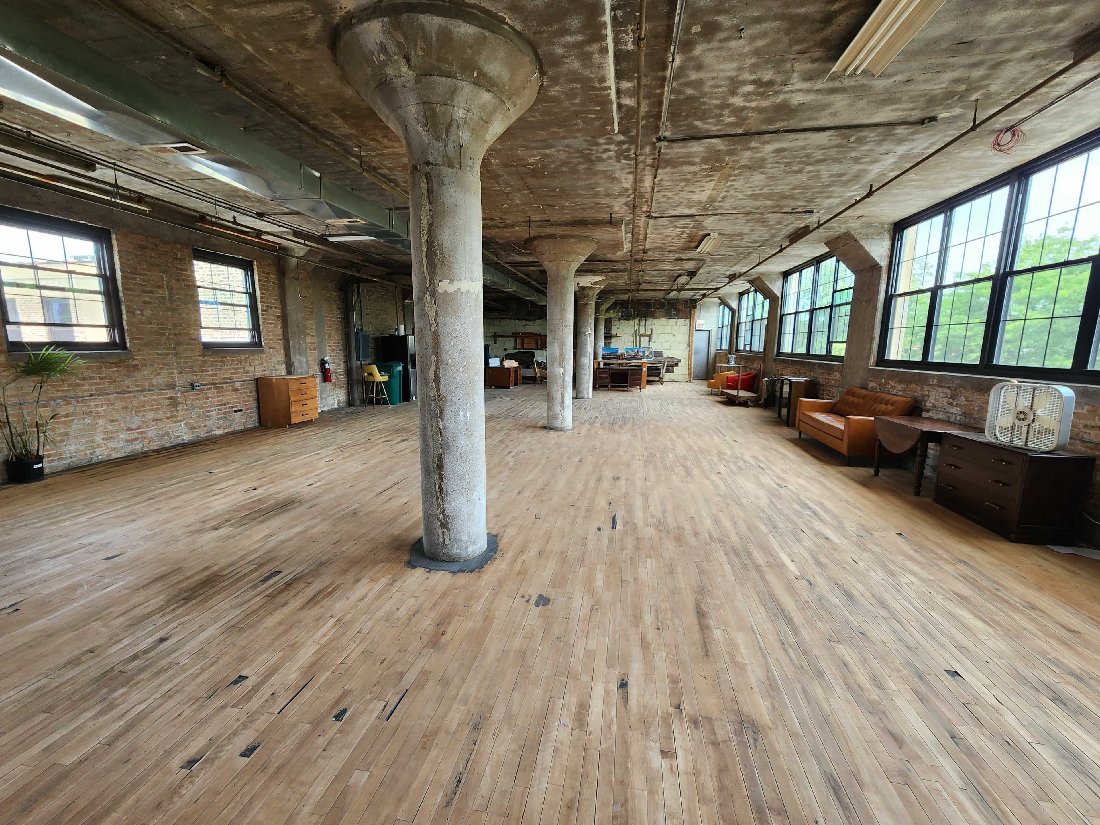 A spacious, industrial-style room with large windows, wooden floors, exposed concrete pillars, and minimal furniture.