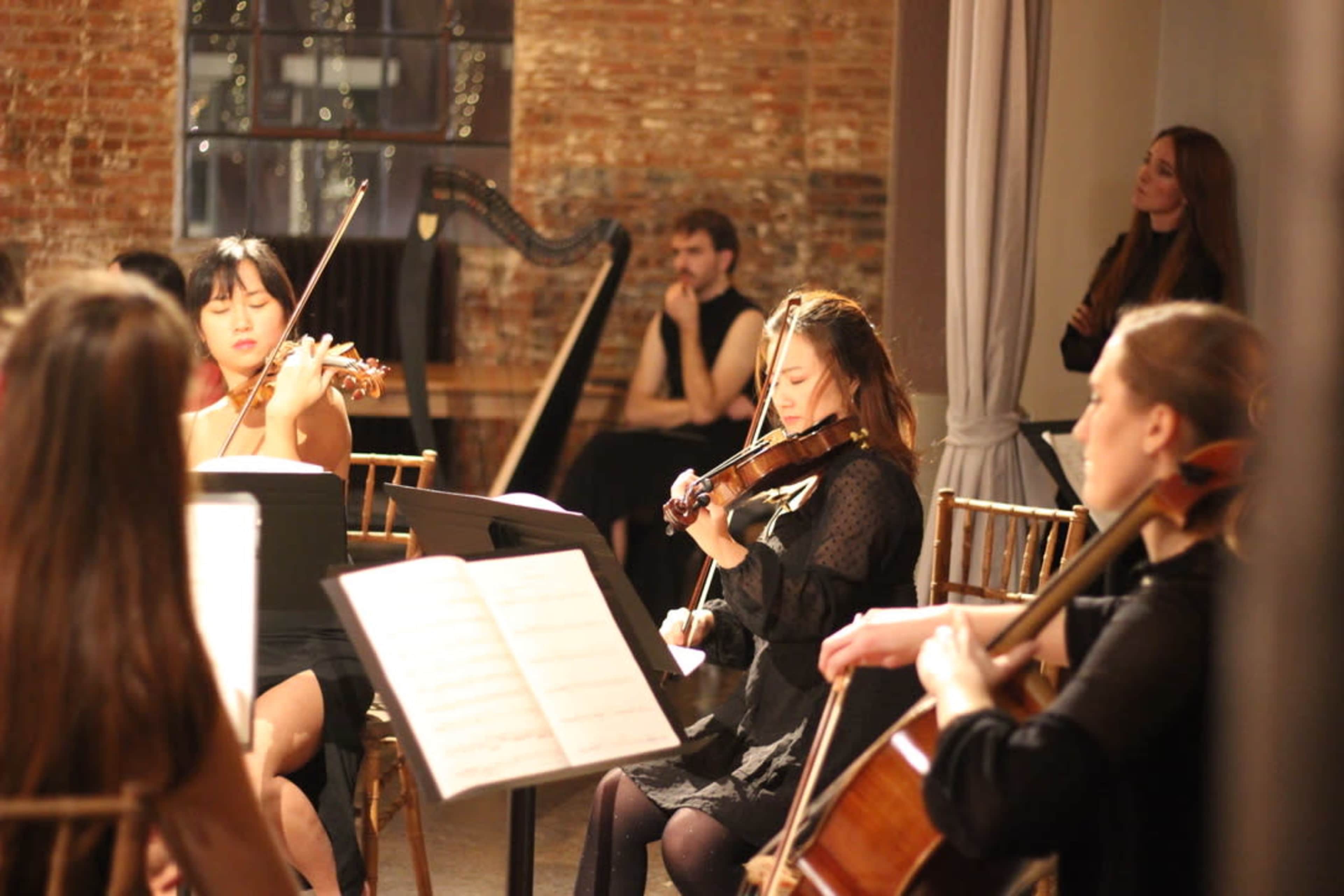 A group of musicians performs together in a dimly lit room, with a harp and various string instruments visible.