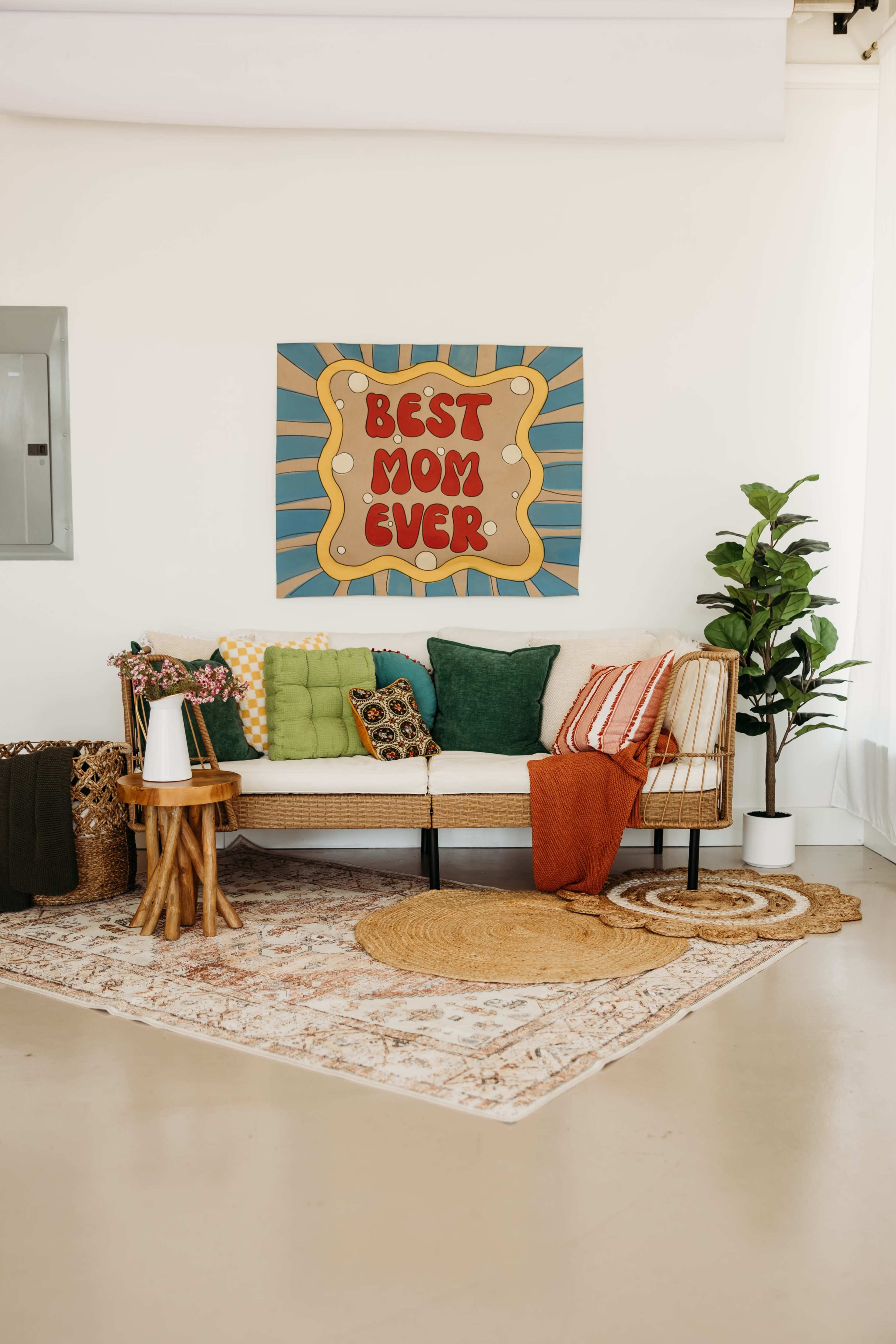 A cozy living room features a rattan couch adorned with colorful pillows, a decorative rug on a light floor, and a large wall art piece that reads "BEST MOM EVER."