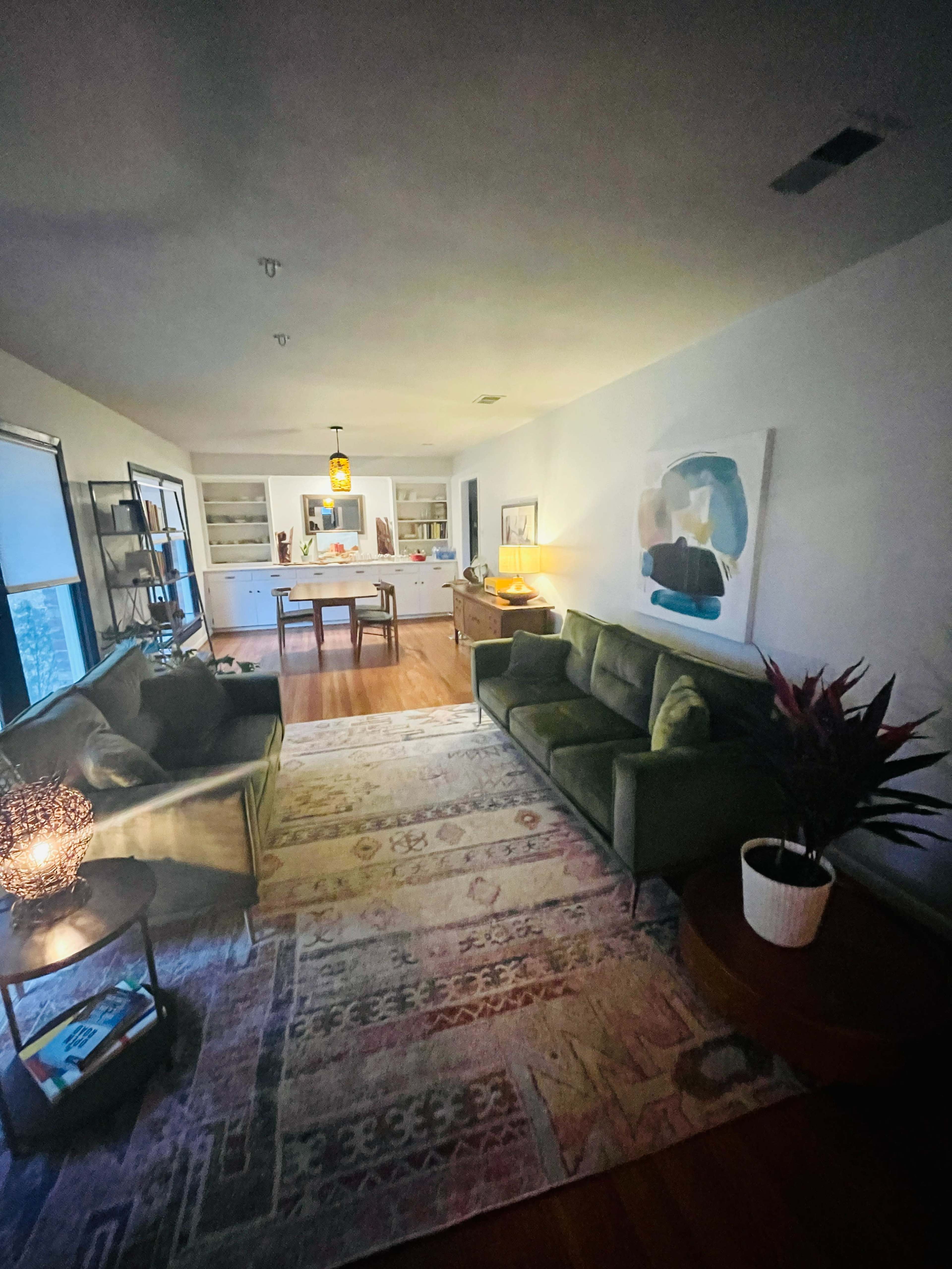 The image shows a living room with a green sofa, a patterned rug, a coffee table, and a dining area visible in the background.