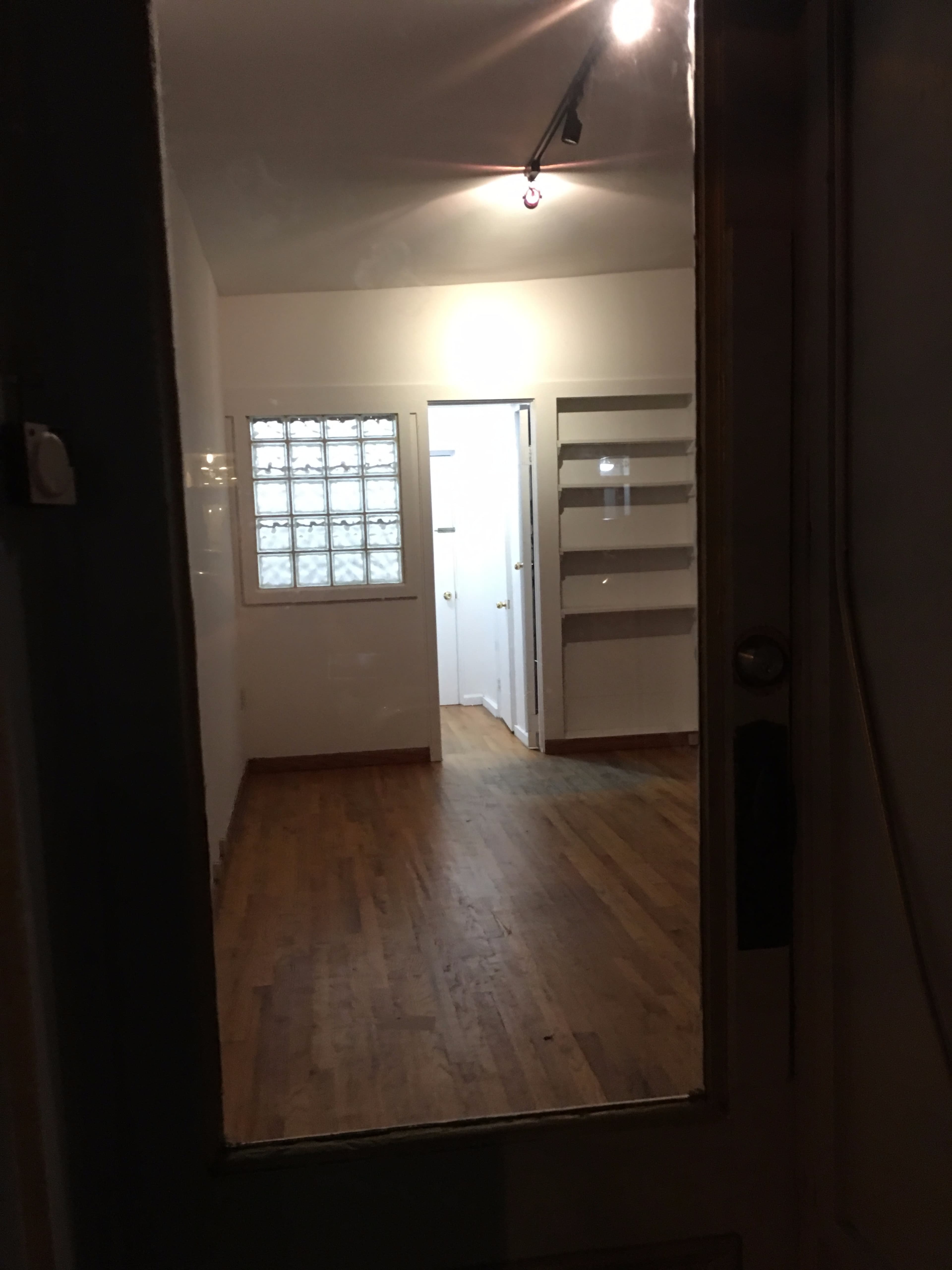 The image shows a wooden-floored room viewed through a glass door, with a hallway leading to another area and a light fixture overhead.