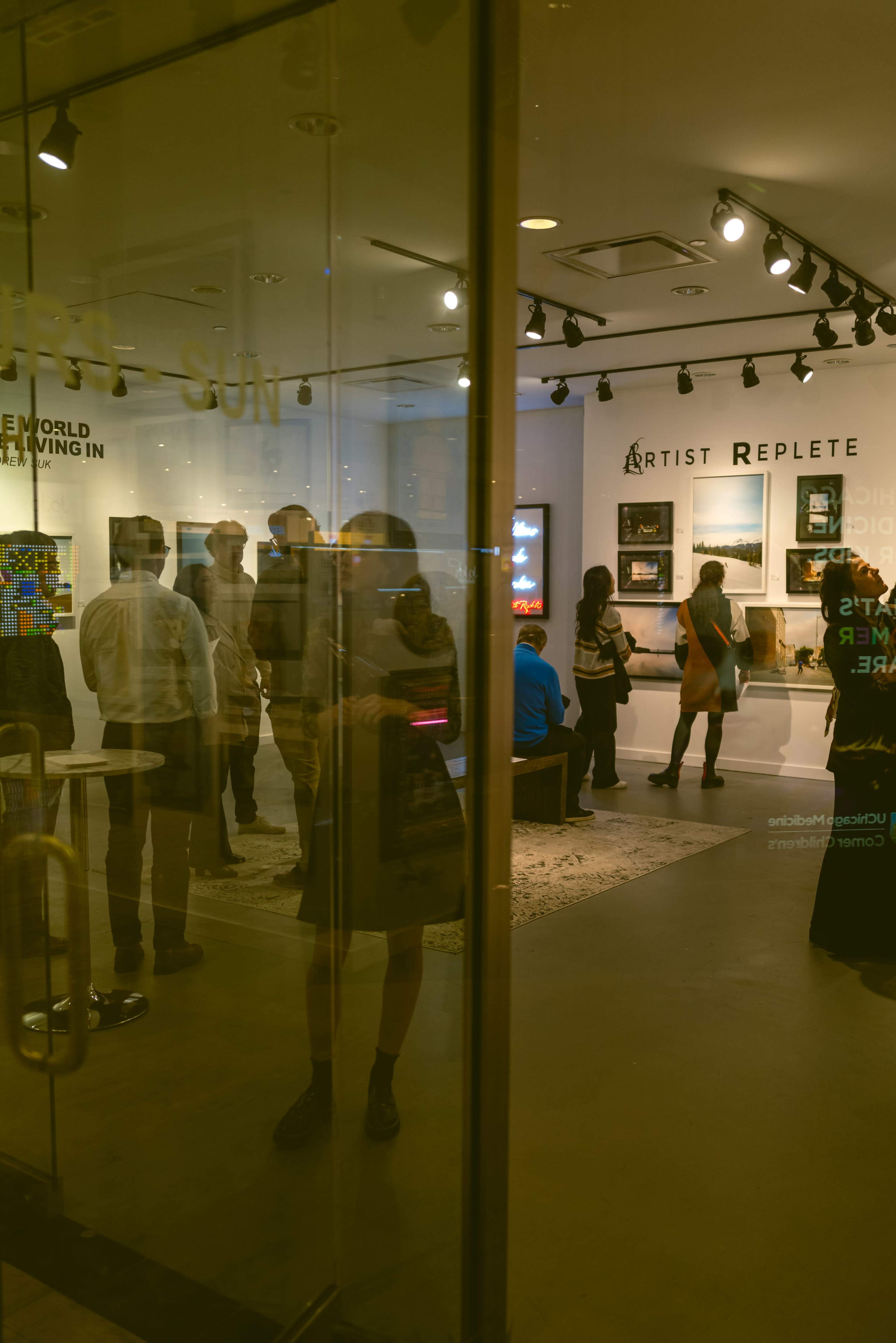 The image shows people exploring an art gallery with framed artworks displayed on the walls and reflections in the glass door.