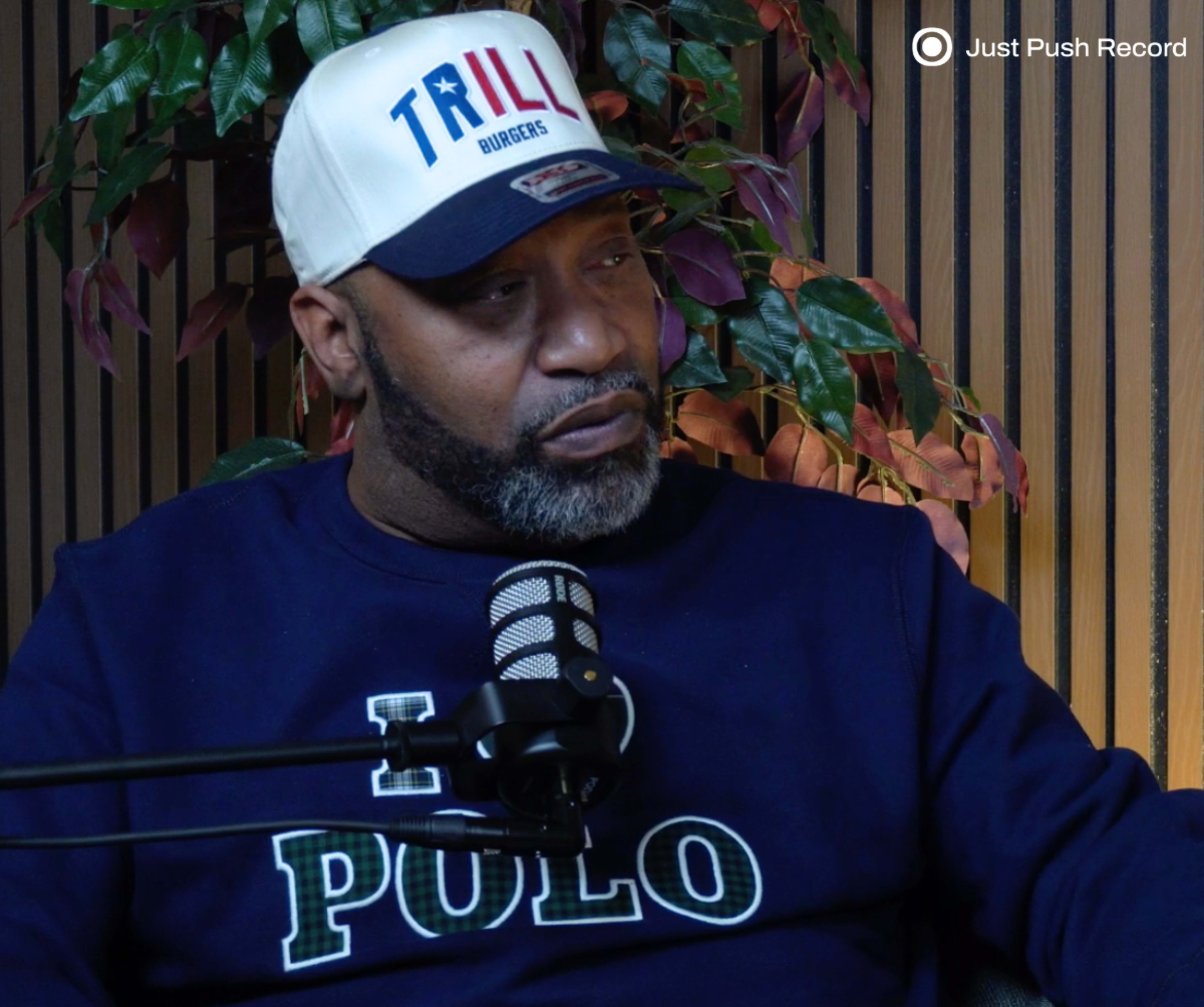 A man in a navy sweatshirt and baseball cap sits in front of a microphone, surrounded by leafy plants and wooden paneling.