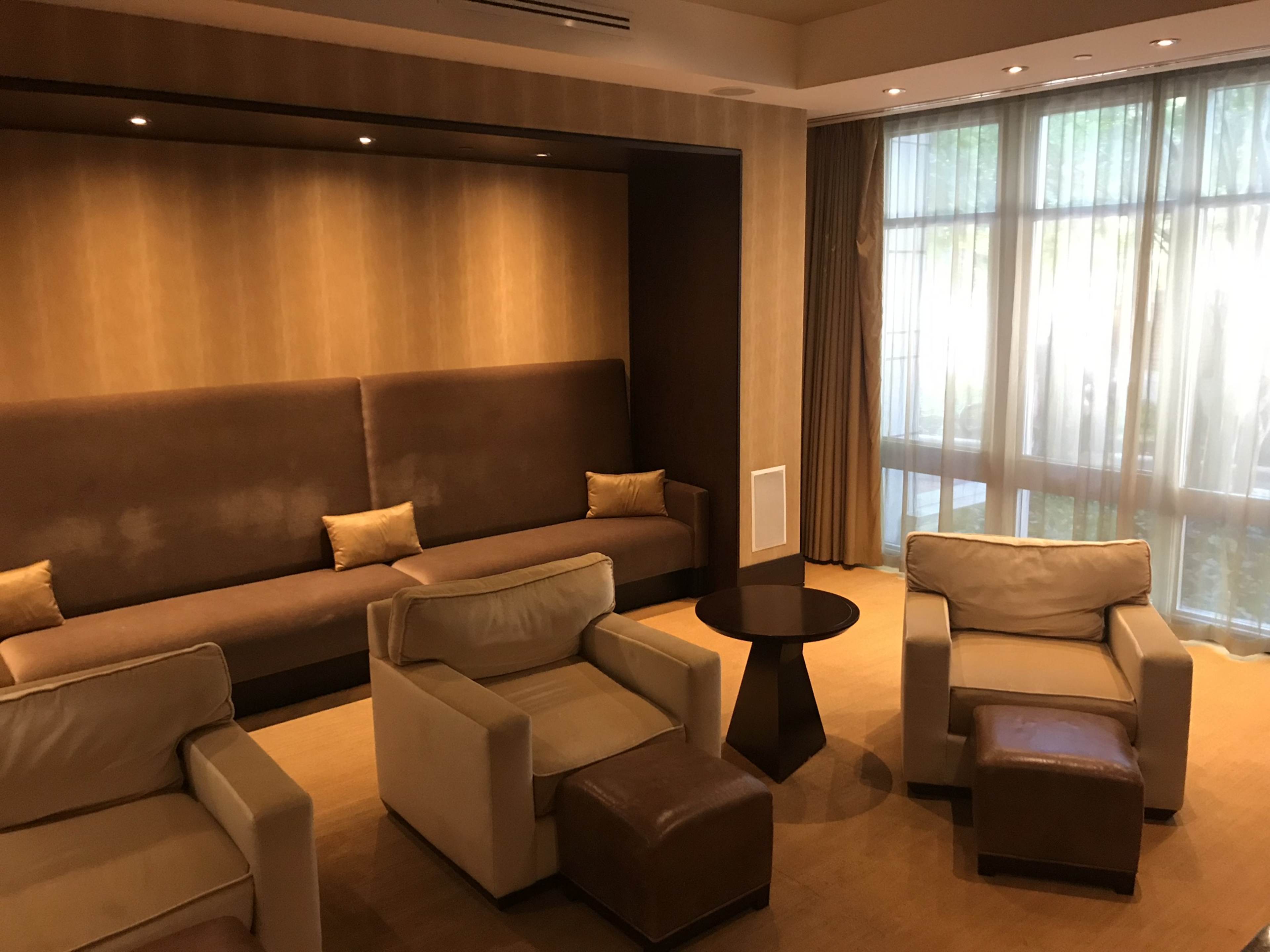 A lounge area with a brown upholstered wall, two light-colored armchairs, a small round table, and a beige sofa positioned against a backdrop of large windows covered by sheer curtains.