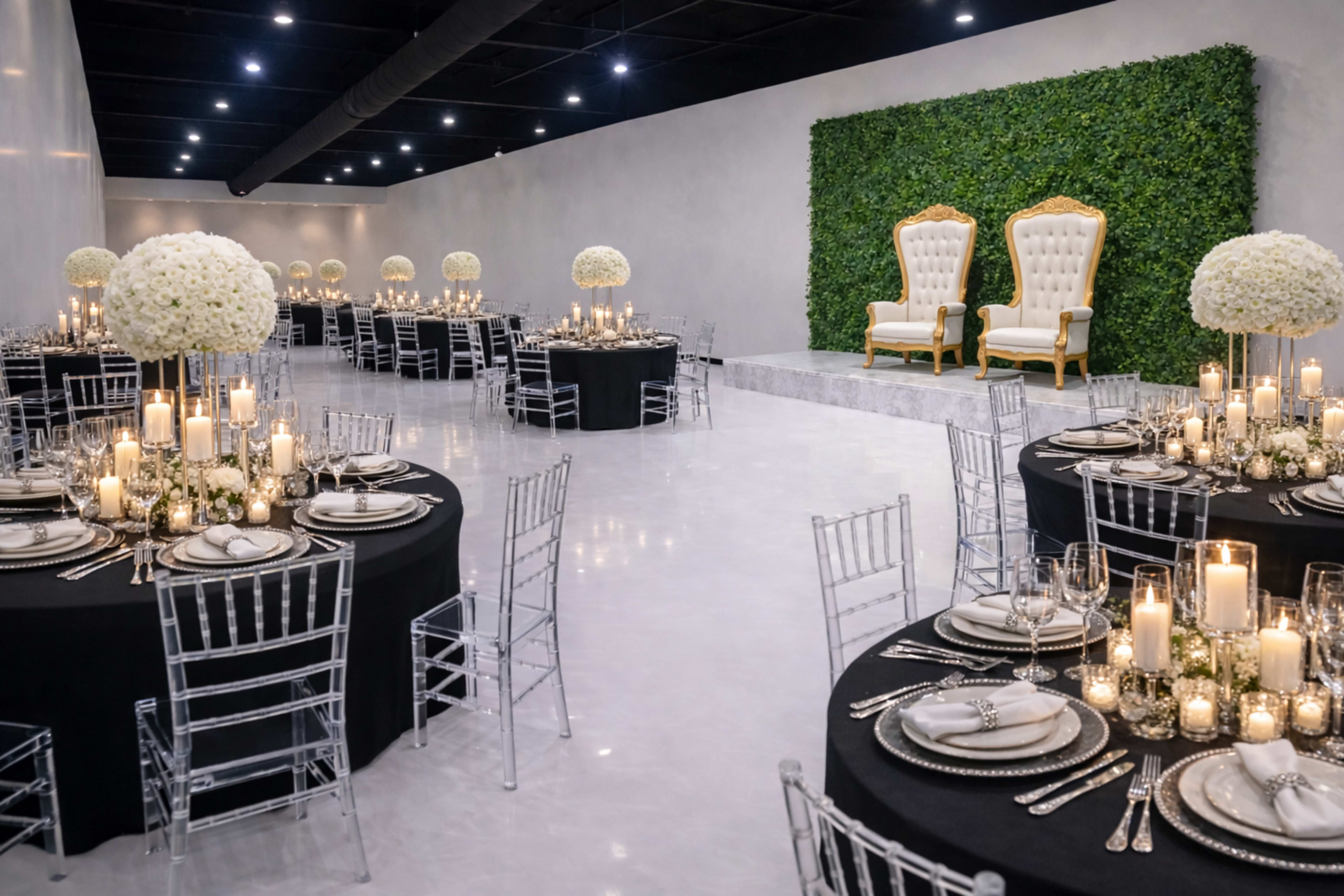The image features an elegantly set banquet hall with round tables adorned with black tablecloths, tall centerpieces of white flowers, and candles, alongside a greenery backdrop and two ornate golden chairs on a raised platform.