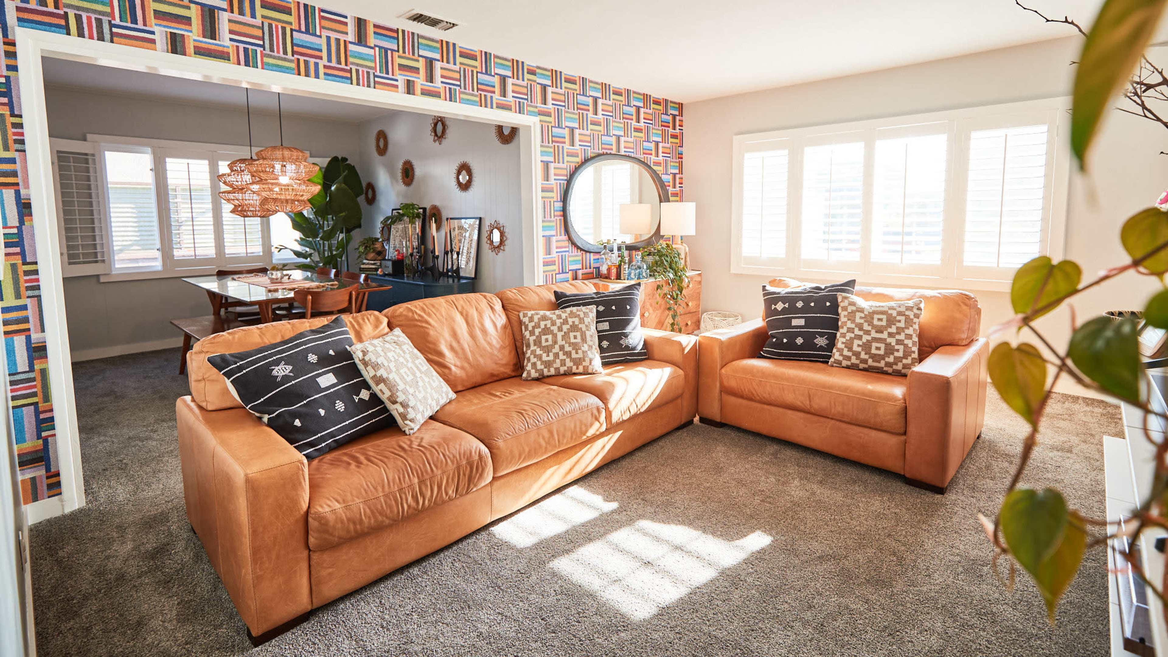 A living room with tan leather sofas, a round mirror on the wall, and colorful patterned wallpaper, illuminated by natural light from the windows.