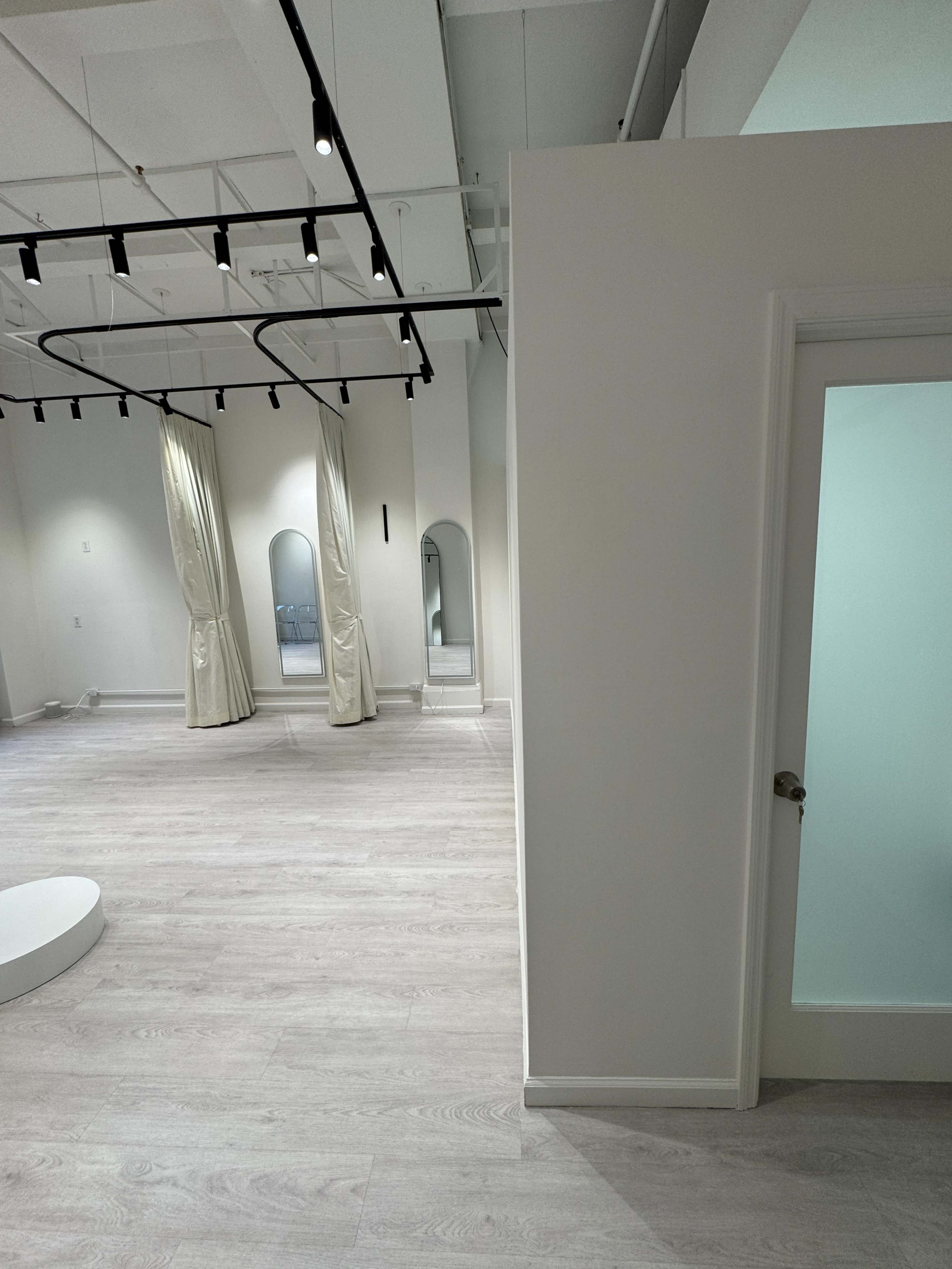 The image shows a spacious, brightly lit interior with light-colored wooden flooring, a partially visible door on the right, and draped fabric sections in the background.