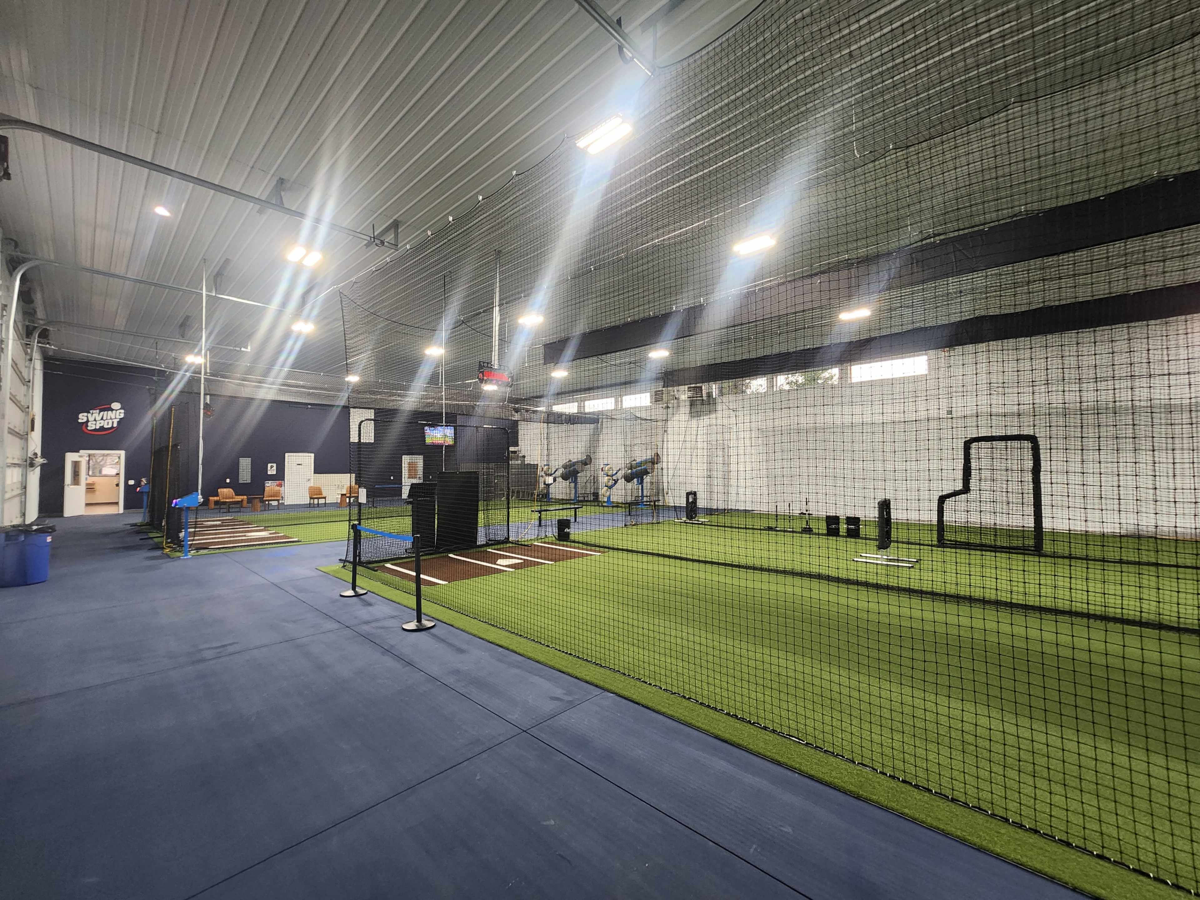 The image shows an indoor sports facility with green turf, batting cages, and training equipment.