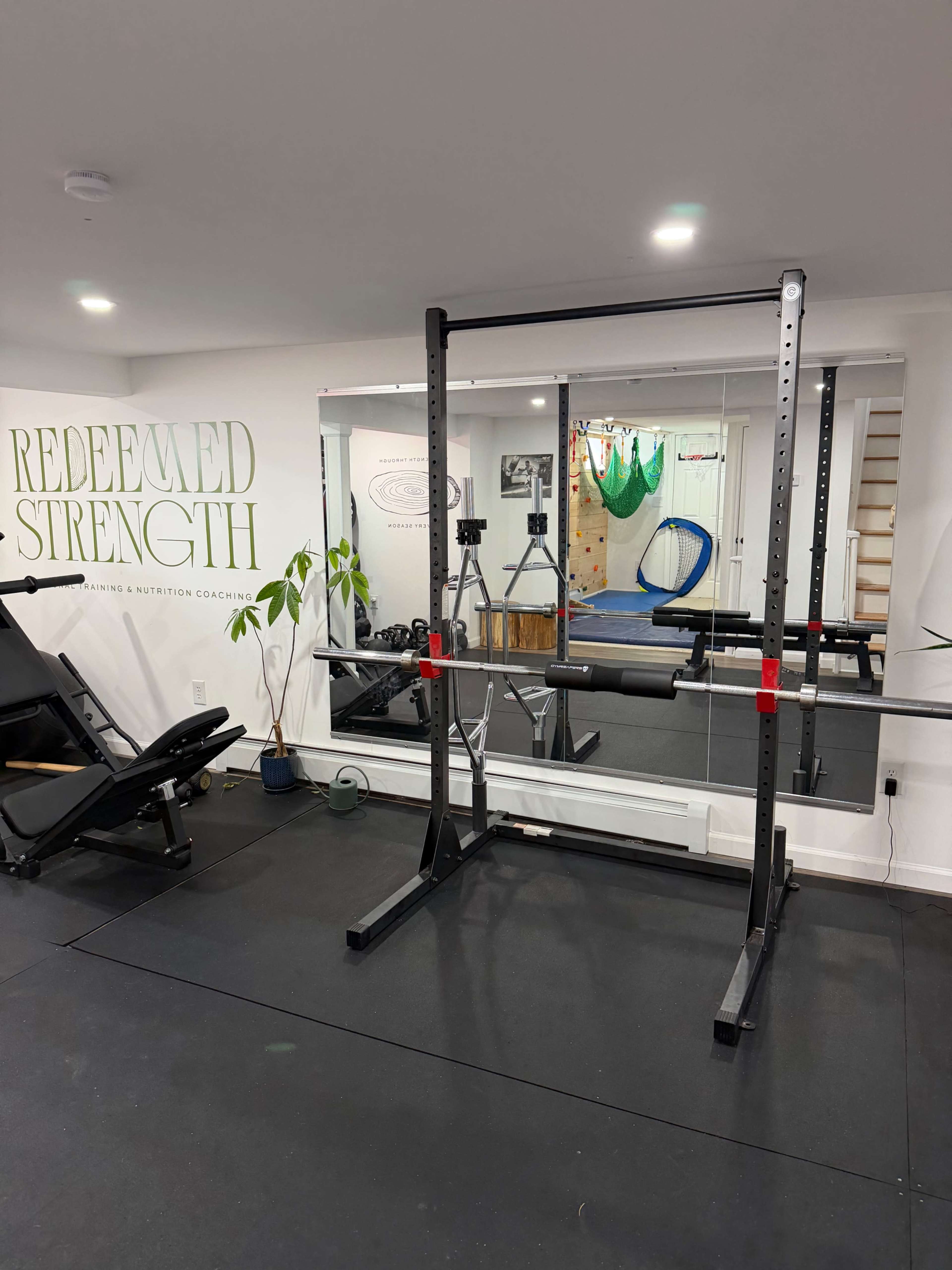 The image shows a gym space with a squat rack, a weight bench, and mirrors on the wall, alongside exercise equipment and greenery.