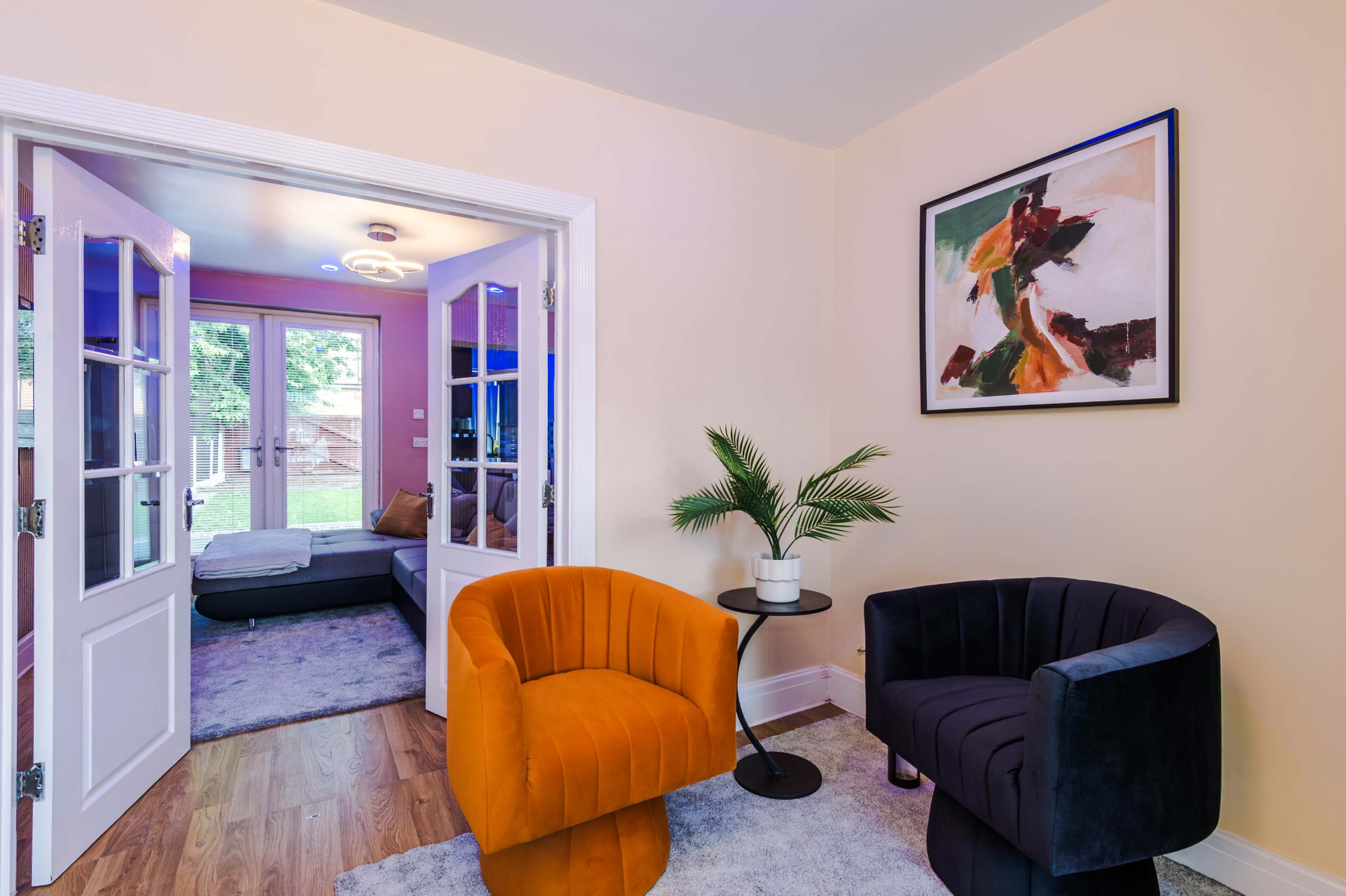 The image shows a cozy room with two accent chairs, a small side table, and an open doorway leading to another area with a couch and large windows.