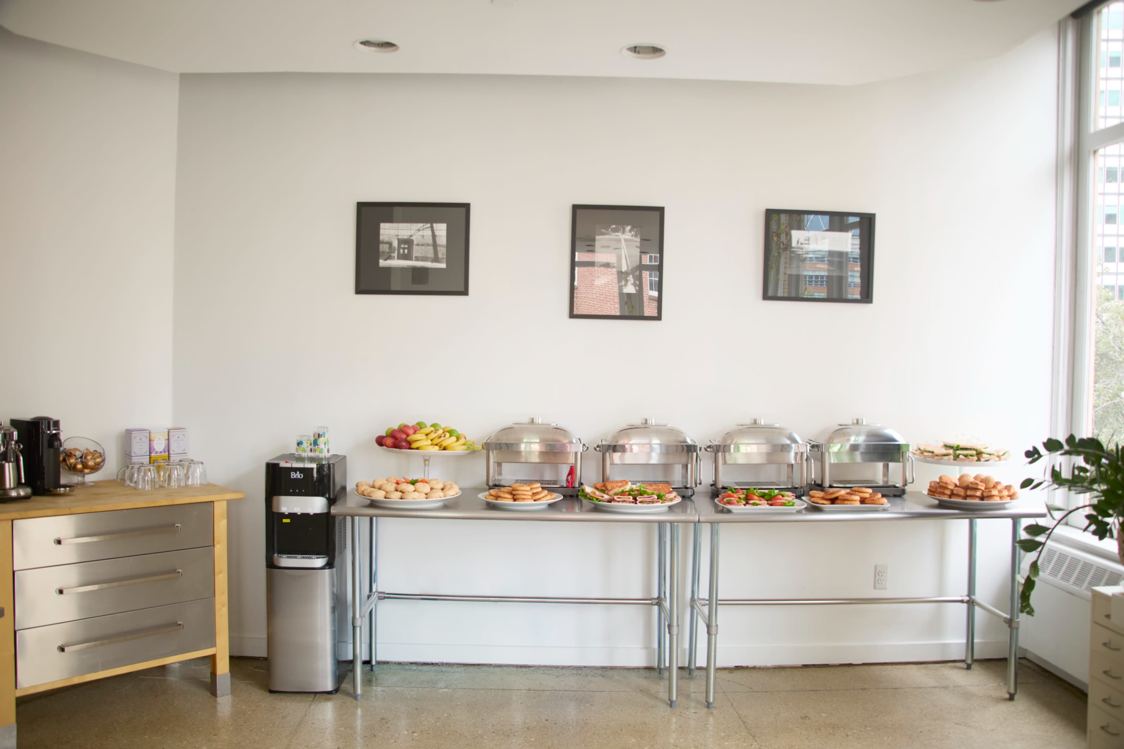 A buffet setup features multiple chafing dishes, a coffee station, and a fruit display against a wall with framed photographs.