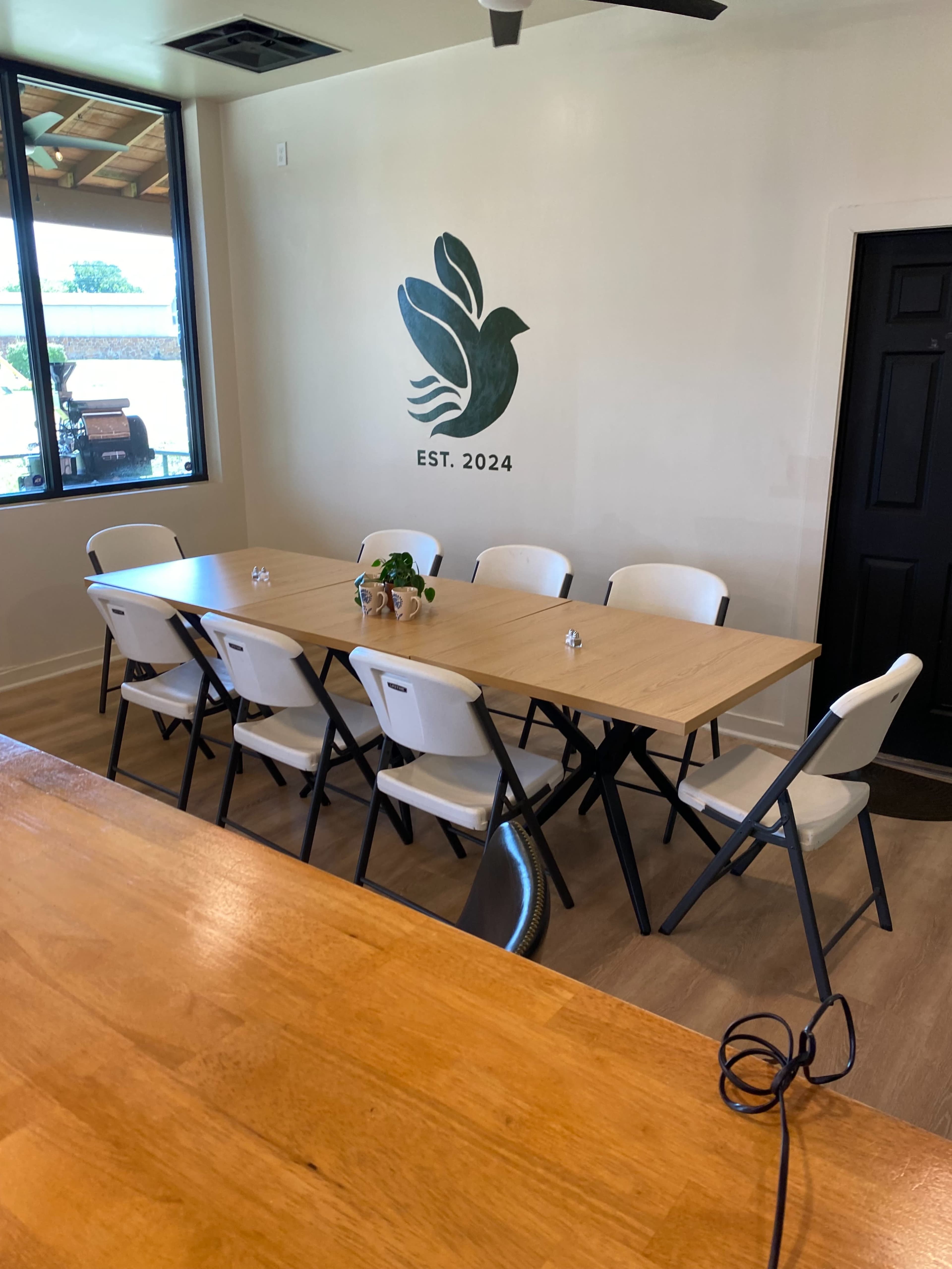 The image shows a spacious room with a long wooden table surrounded by eight folding chairs, and a wall featuring a green bird design and the text "EST. 2024."