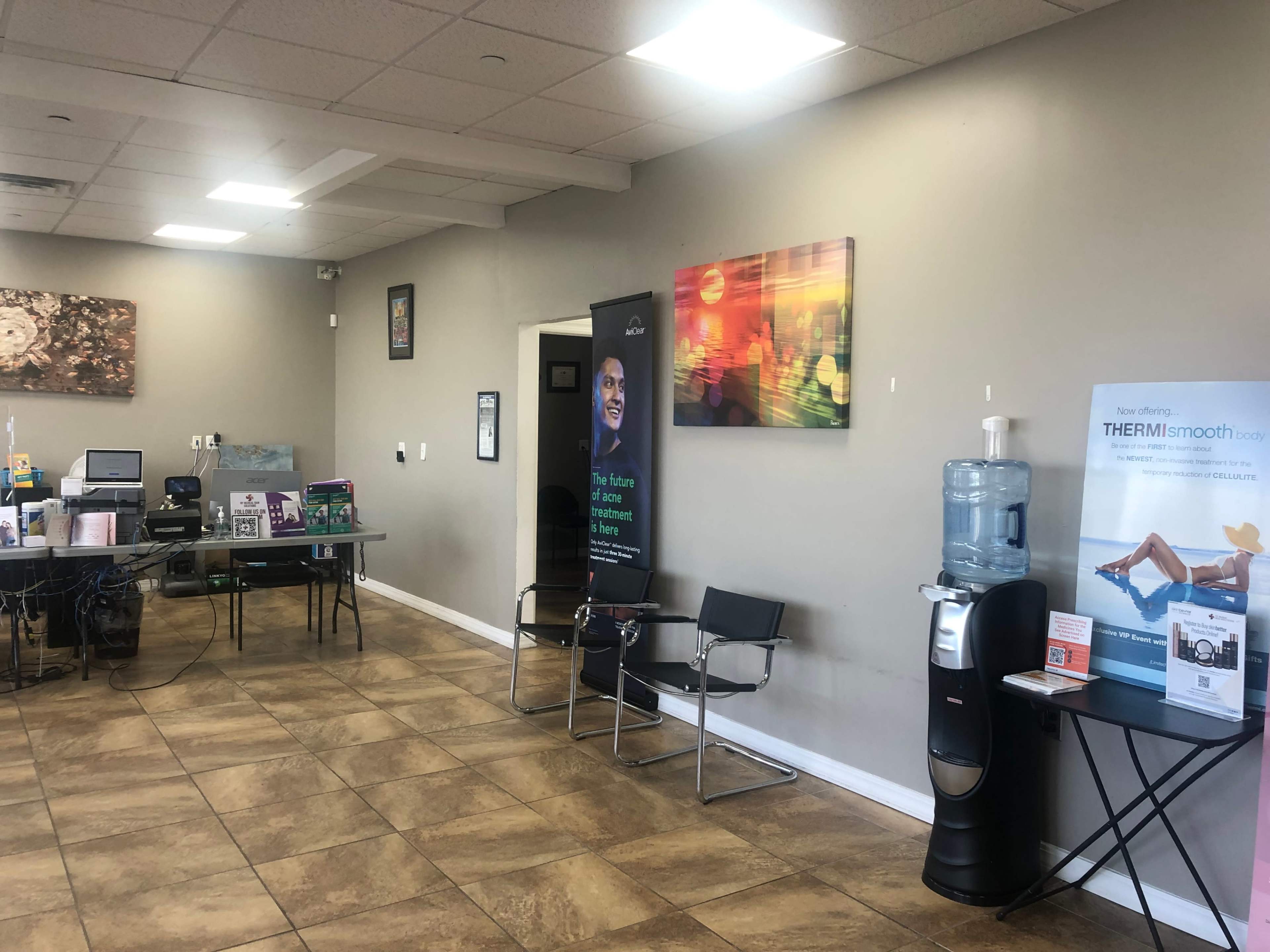 The image shows an interior space with a reception area featuring a water cooler, chairs, a table with promotional materials, and colorful artwork on the walls.
