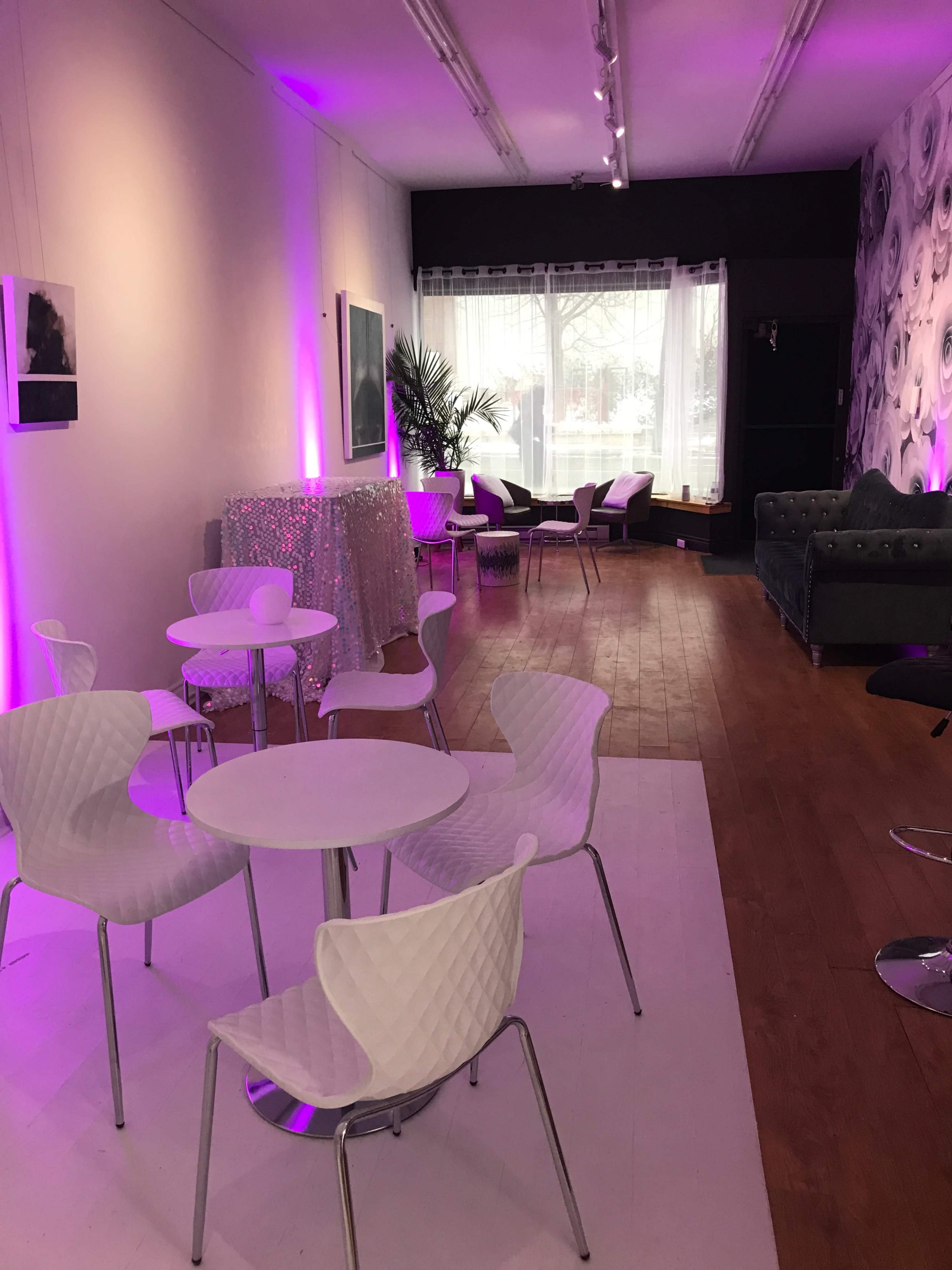 A stylish interior space with white tables and chairs, purple lighting, and a mix of seating areas, including a bar and a plush couch.
