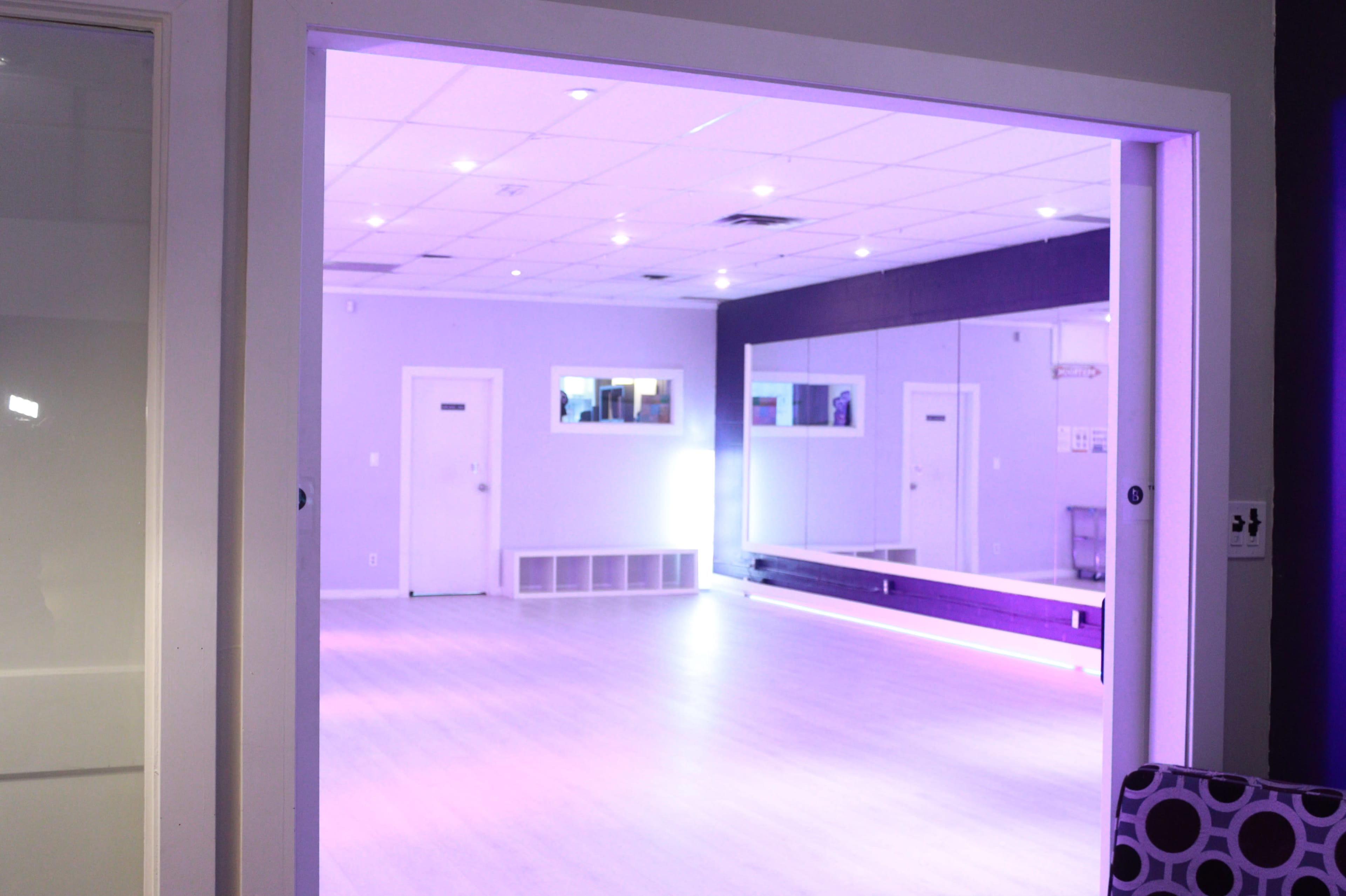 A dance studio with purple lighting features a mirrored wall and a spacious wooden floor.