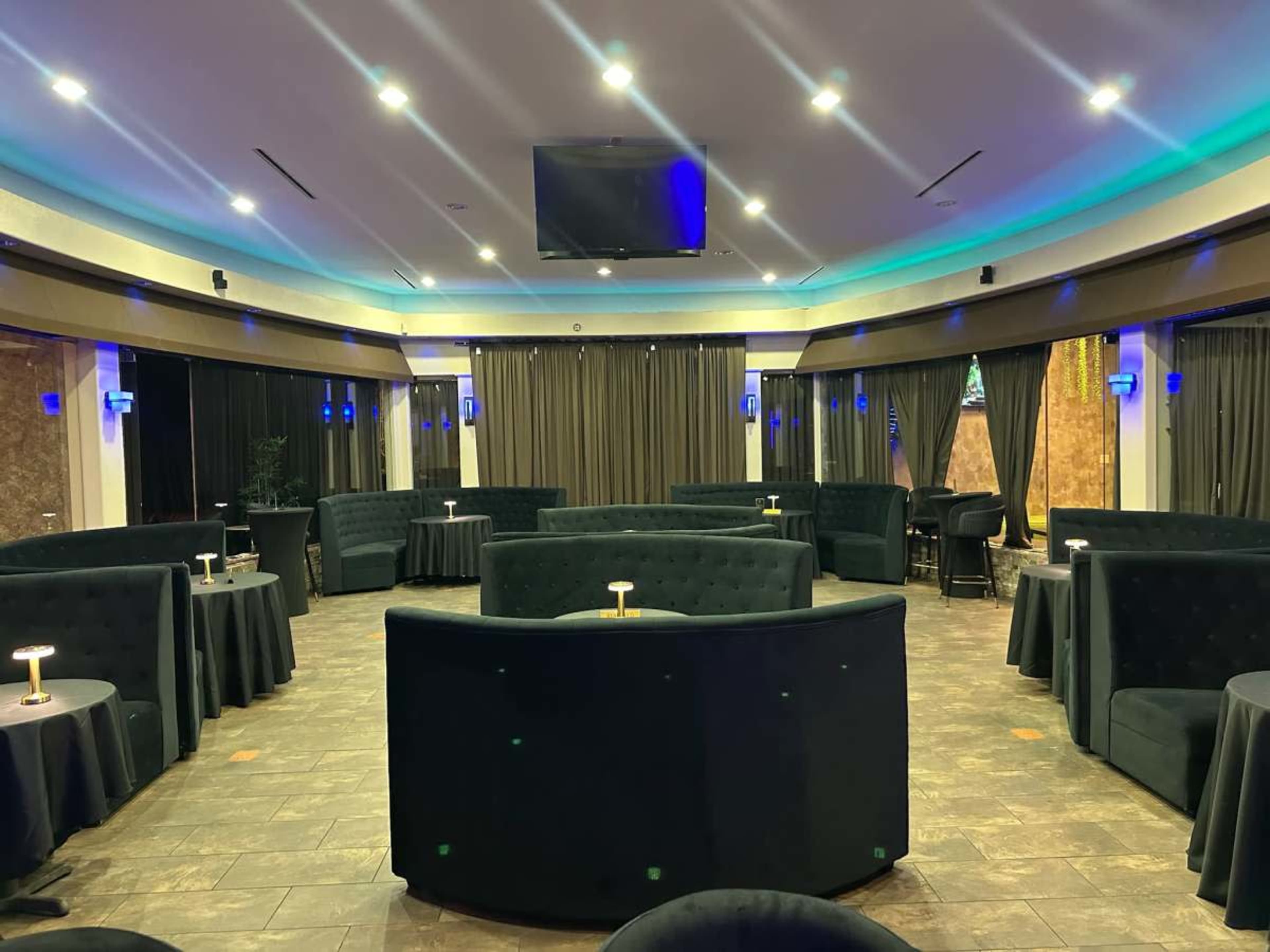 The image shows a modern lounge with dark seating arrangements and round tables, illuminated by soft lights and curtains along the walls.